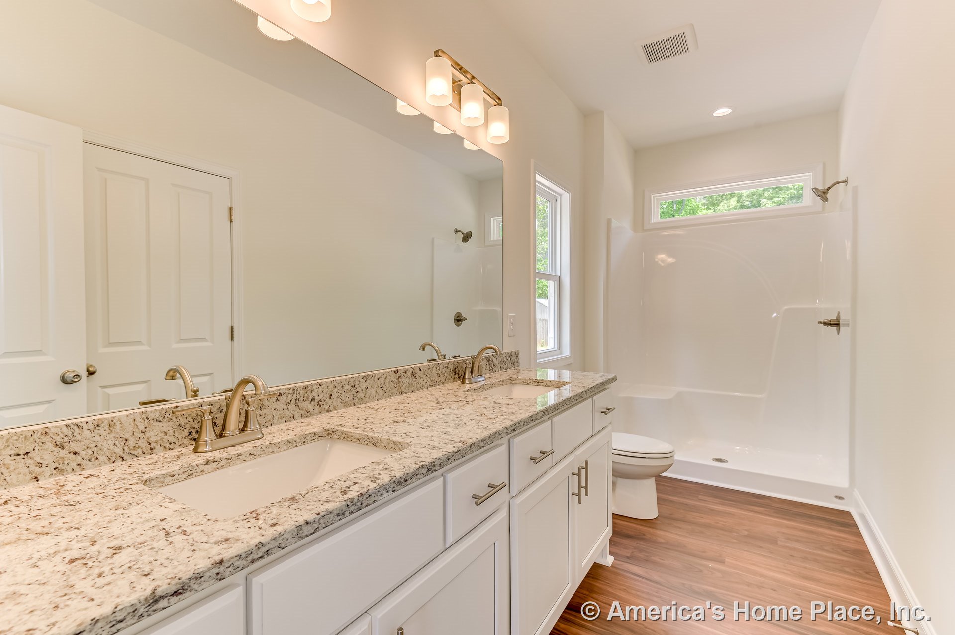 Double-sink vanity with granite countertop, white shaker cabinets, and wall-mounted lighting; walk-in shower enclosure with glass door; wood-look flooring, large mirror, recessed