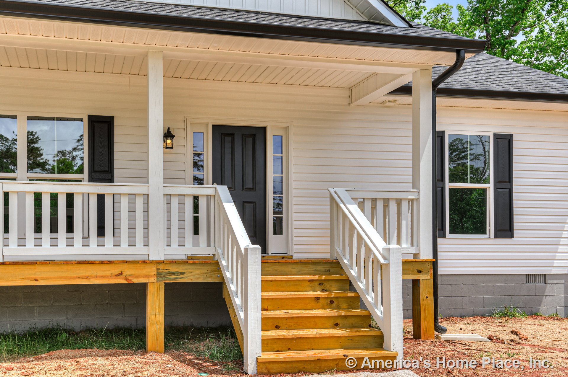 Covered front porch with white railings and wooden steps, black front door, white vinyl siding, black window shutters, exterior wall-mounted light, gabled roof, and concrete block
