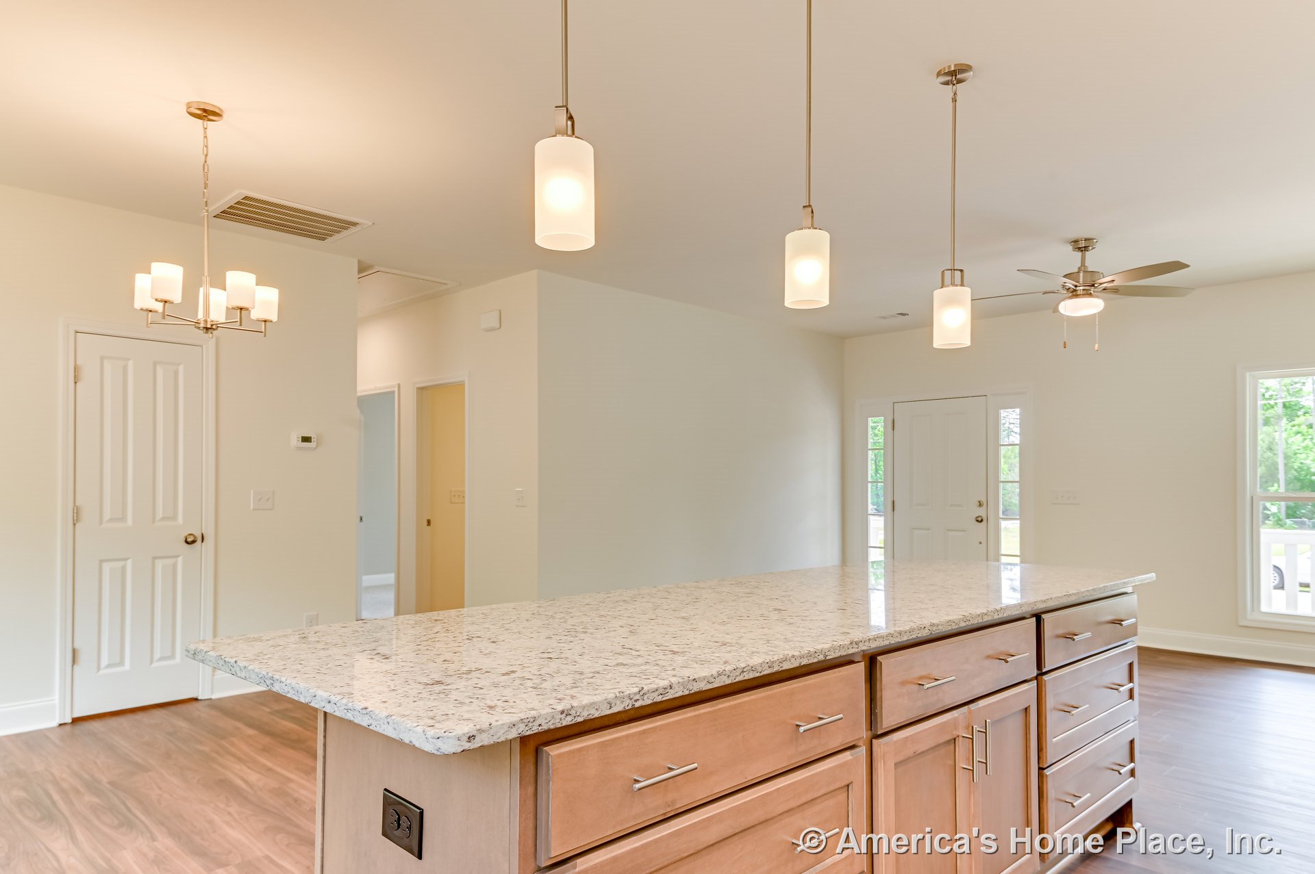 Spacious kitchen with large granite island, wood cabinetry with metal pulls, pendant lights above island, open floor plan extending to living area with ceiling fan, multiple