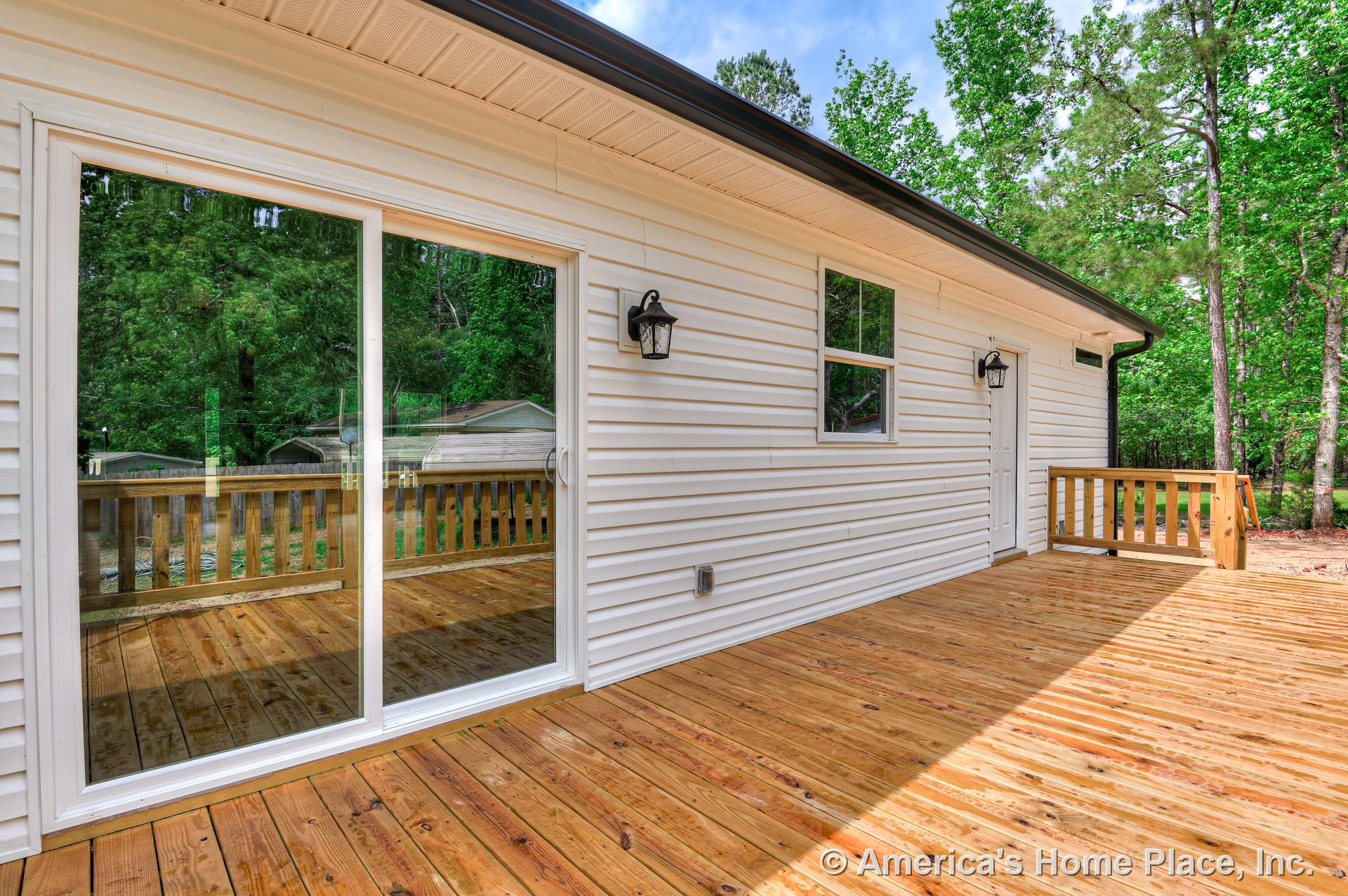 Spacious wooden deck with matching railing, attached to home with white vinyl siding, sliding glass door, single exterior window, and wall-mounted lantern lights.