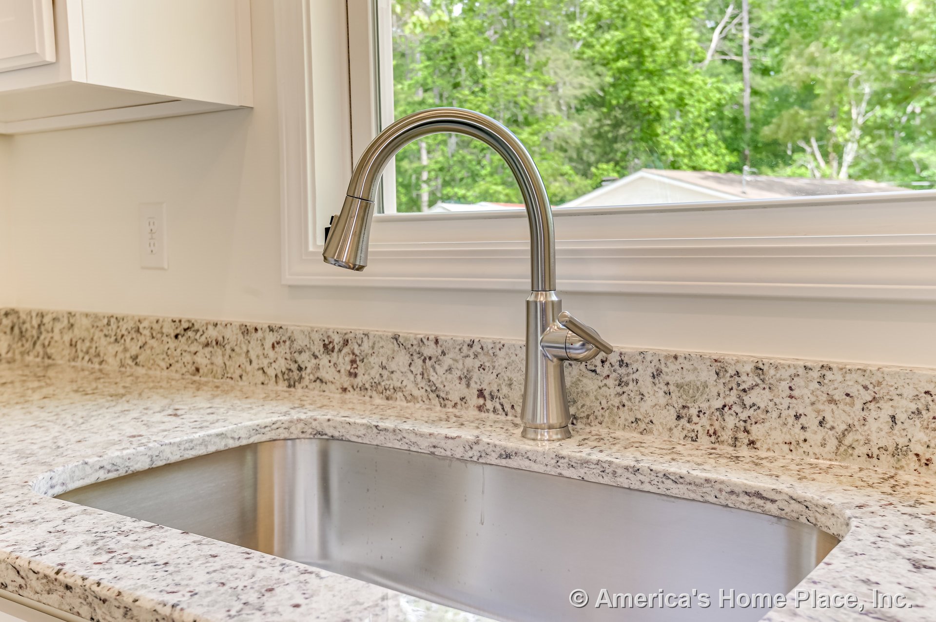 Stainless steel undermount sink with brushed nickel faucet set in speckled granite countertop, white cabinetry below, and large window with detailed trim above allowing natural