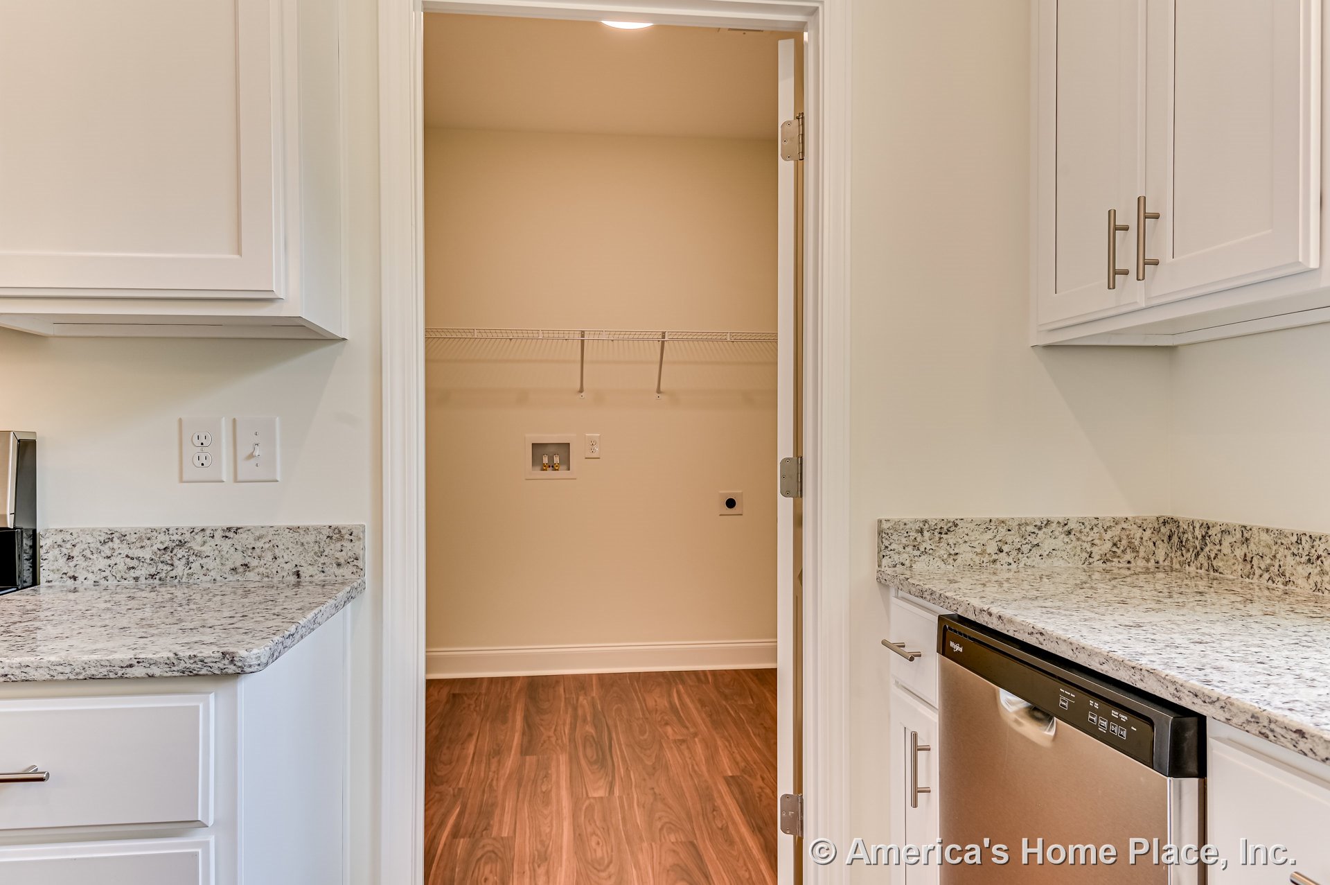 Granite countertops and white cabinetry in kitchen with stainless dishwasher, open doorway to laundry room featuring wood-look flooring and wall-mounted wire shelving.