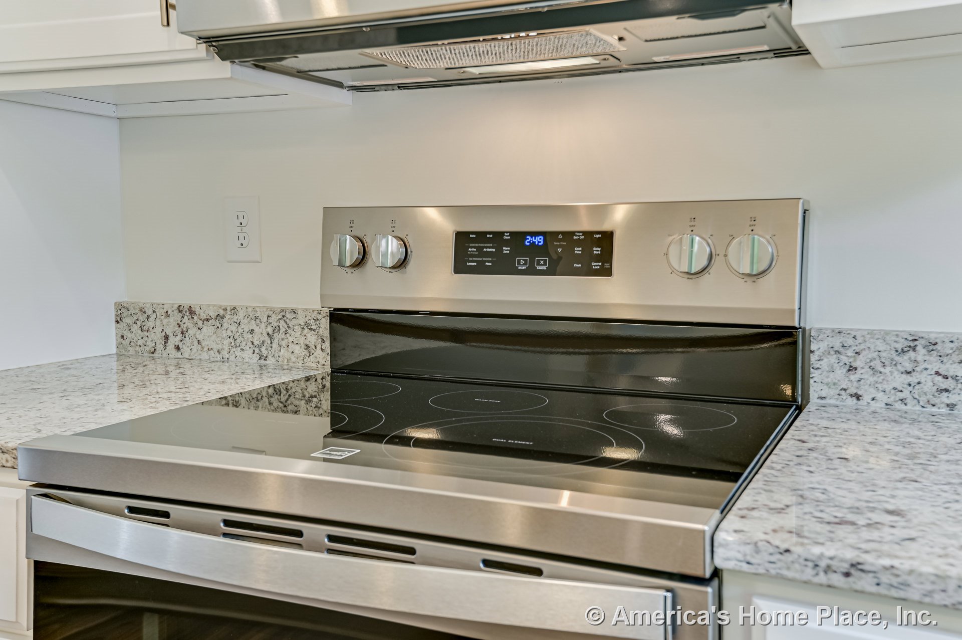 Stainless steel electric range with smooth cooktop and digital controls set between granite countertops and matching backsplash, white cabinetry above, built-in range hood, and