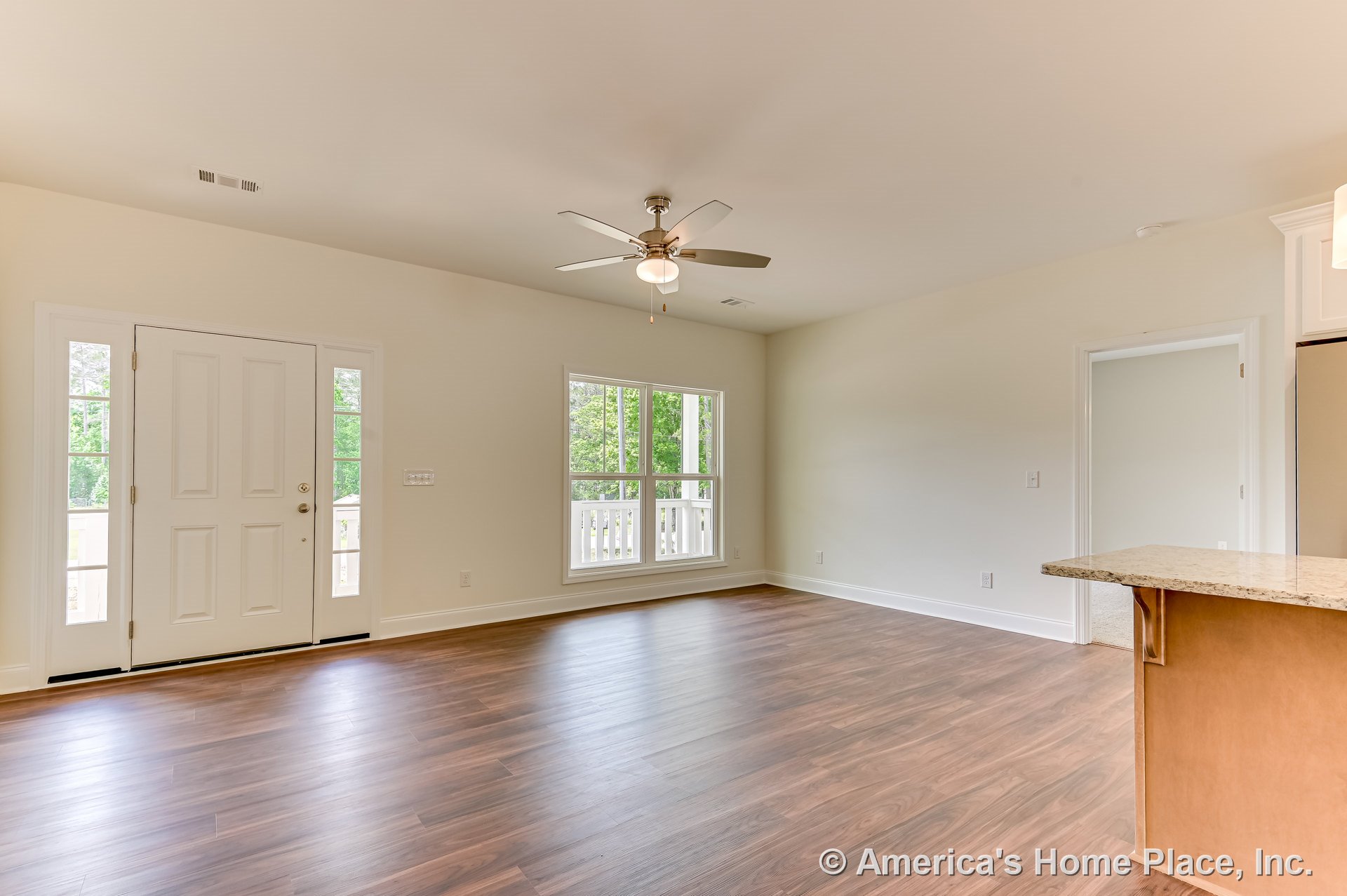 Open living area with wood-look plank flooring, ceiling fan with integrated light, large front windows and entry door with sidelights, white trim and baseboards, granite kitchen