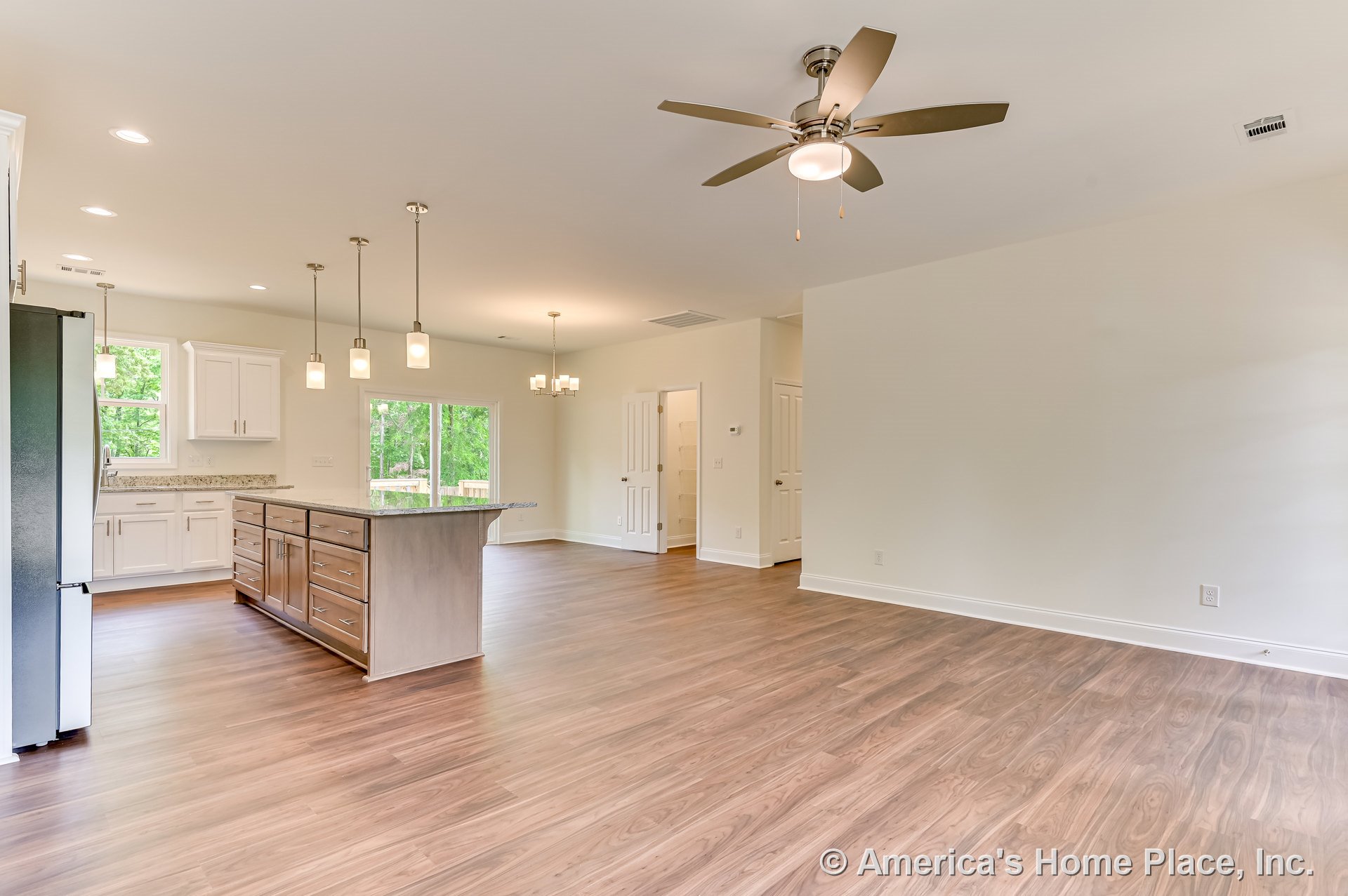 Open-concept kitchen and living area with wood-look flooring, white upper and lower cabinets, granite countertops, kitchen island with drawers, large windows, recessed and pendant