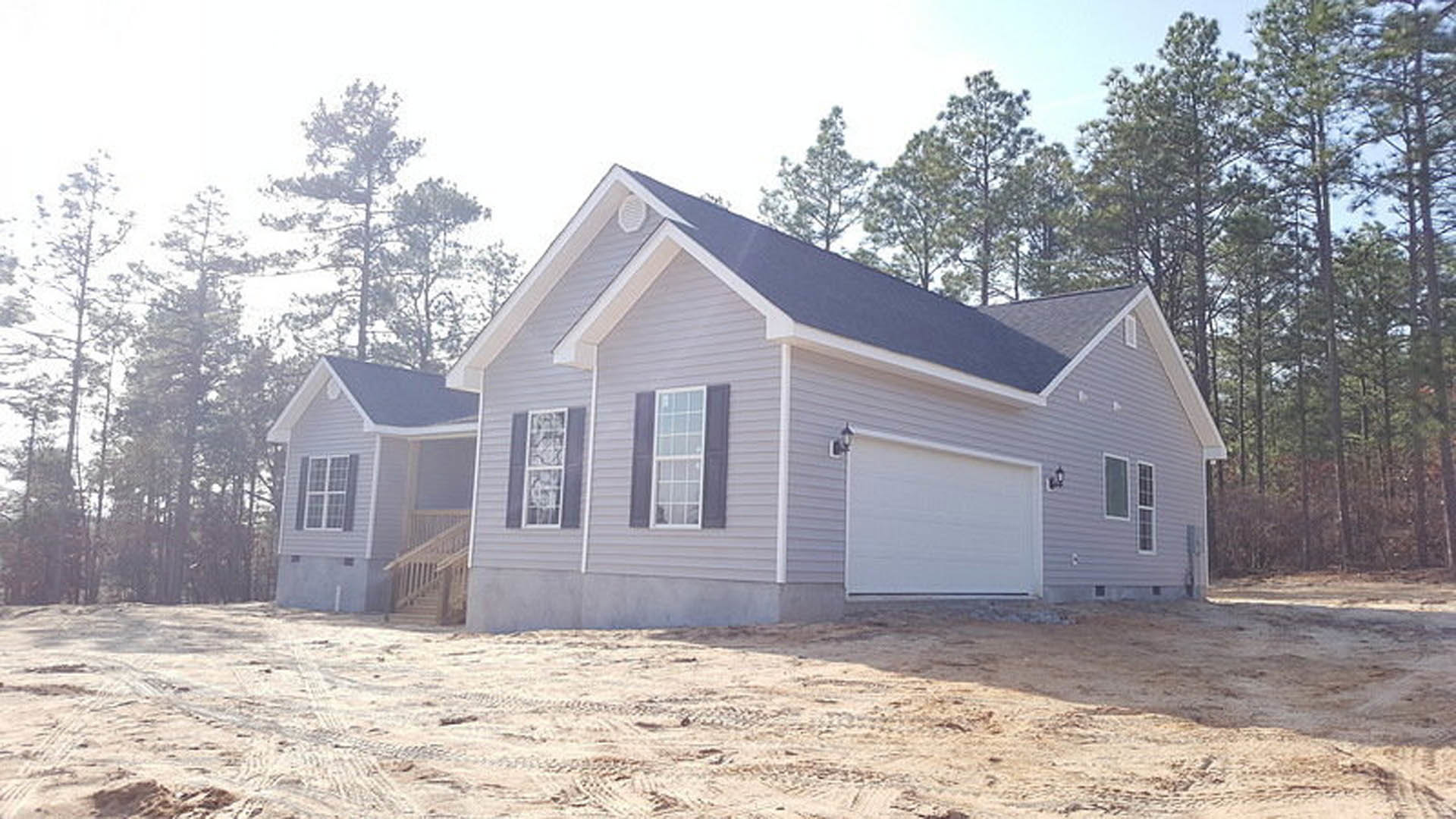 Grey house under construction with attached garage, white roller shutter, white-framed window, wooden railing, dirt foreground, and trees in the background