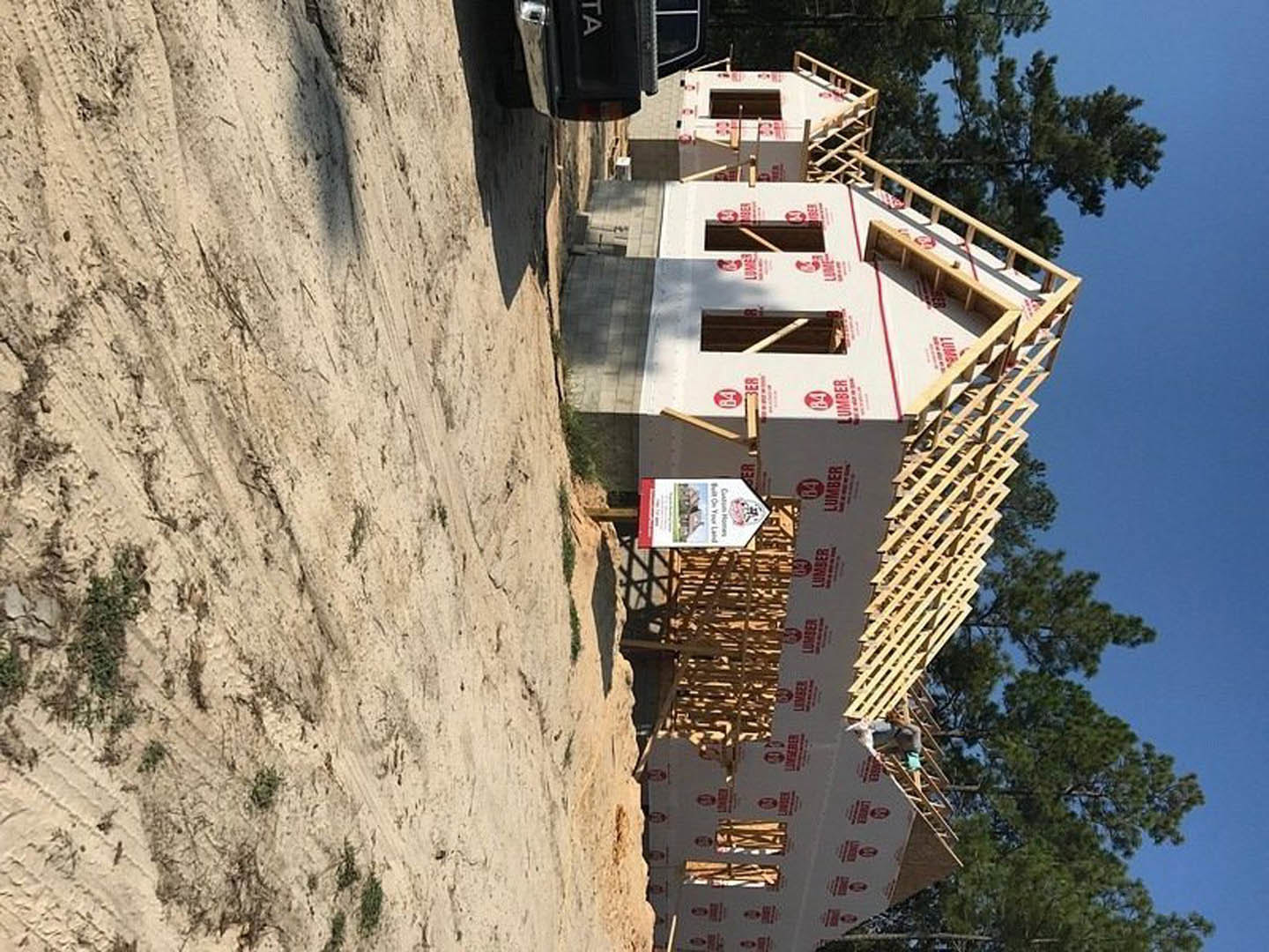 Two-story house framed with exposed wood and sheathing, construction truck parked in front, surrounded by tall trees and dirt ground