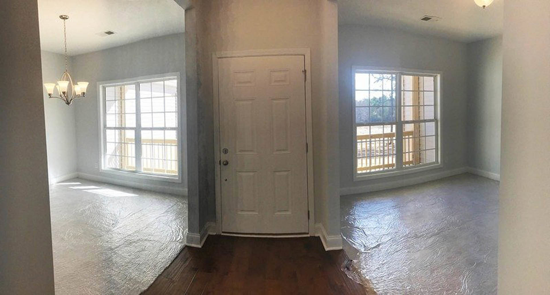 Bright room with white paneled door, two large windows overlooking deck and trees, hanging chain light fixture, smooth plaster walls, and hardwood flooring