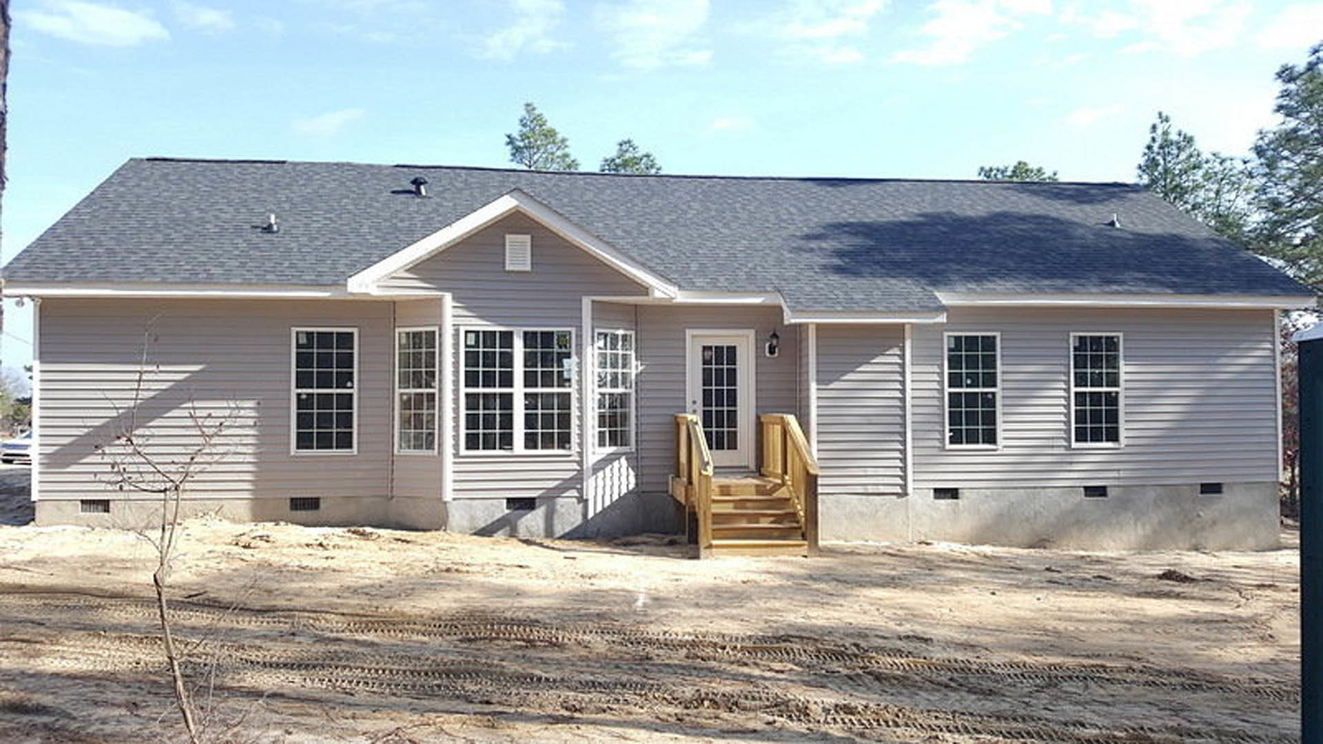 Two-story home under construction with wood siding, porch, exterior stairs, square-paned windows, dirt path with tire tracks, and surrounding trees under a cloudy sky