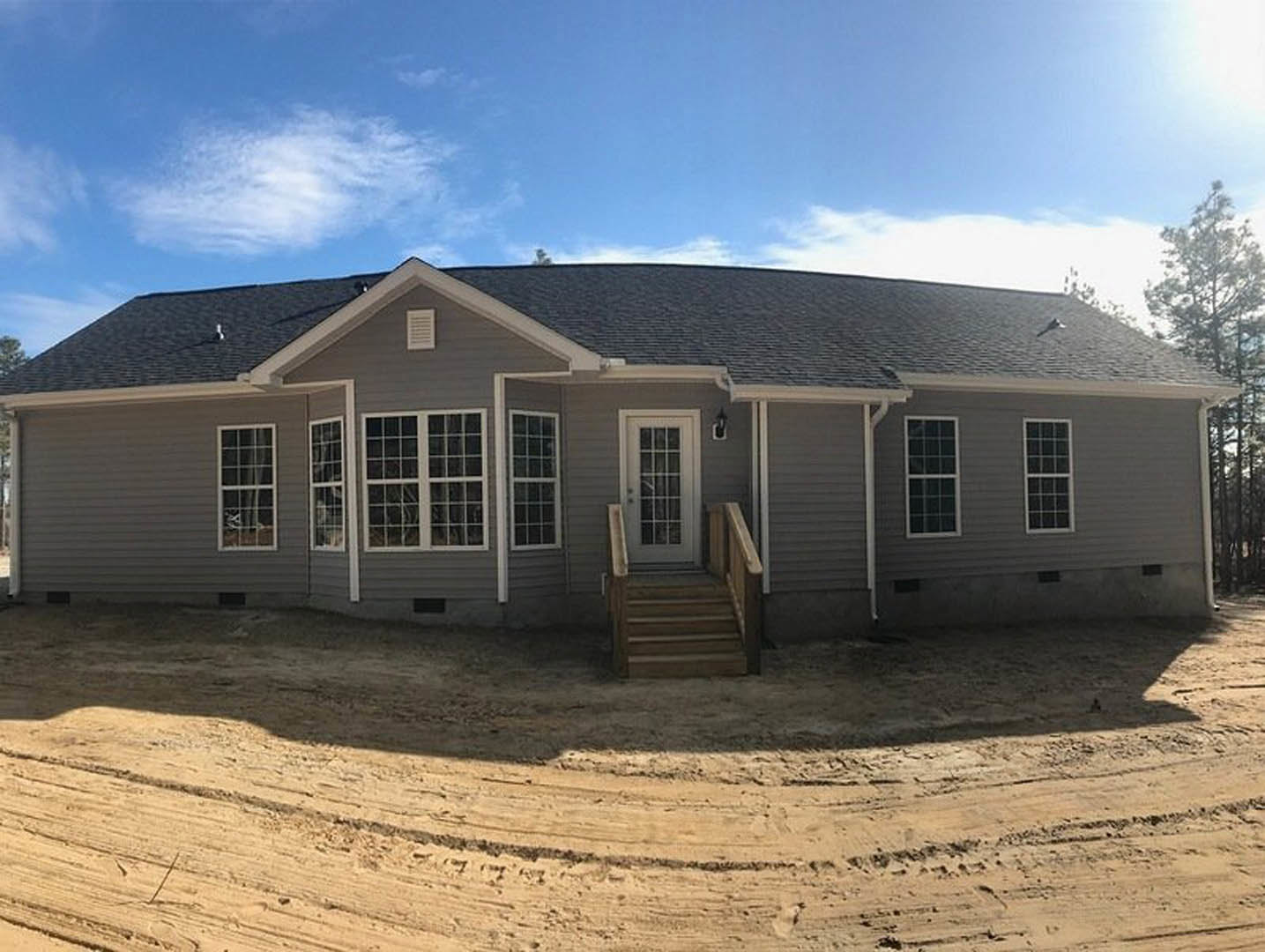 Two-story house with wooden porch and stairs, glass-paneled front door, dirt road with tire tracks, surrounded by trees and cloudy sky
