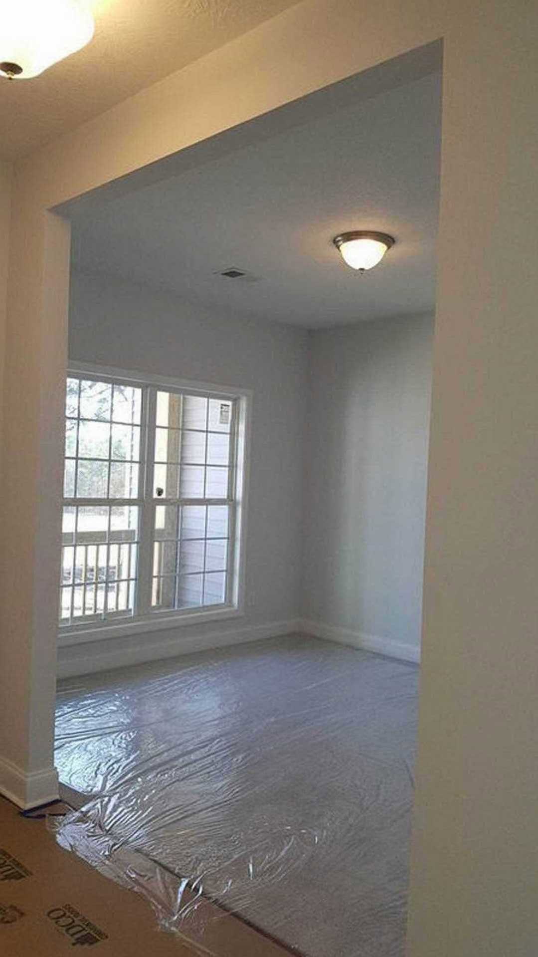 Bright room with large window, white door with glass panes, ceiling light fixture, plastic wrap covering light-colored floor, smooth plaster walls.