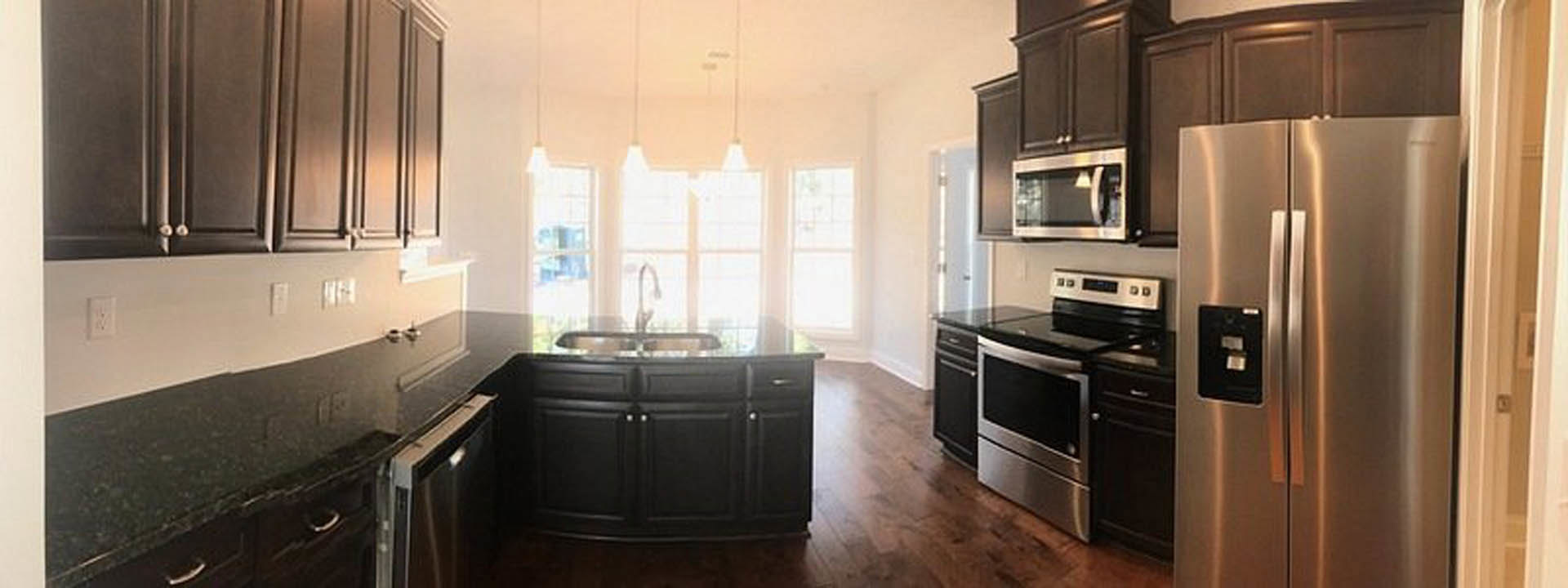 Black kitchen cabinets with brushed metal hardware, stainless steel appliances, wood plank flooring, white quartz countertops, and a large window letting in natural light