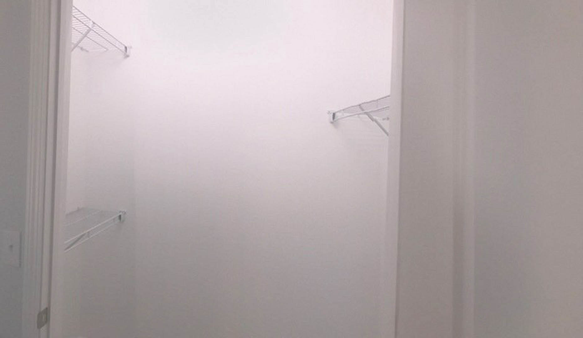 White-painted room featuring a white wall, white door, built-in white shelves with metal rods, electrical outlet, and a person standing on a bench.