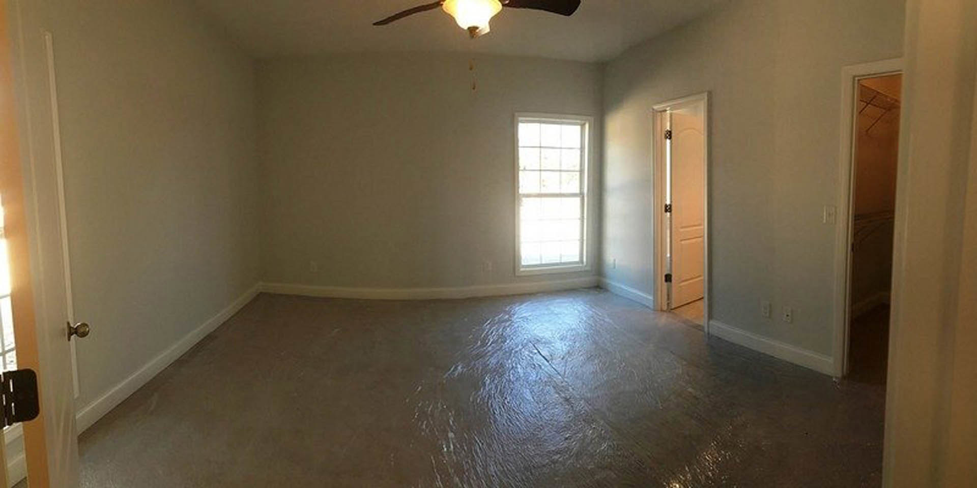 Neutral-toned room with laminate flooring, plaster walls, ceiling fan, large window letting in natural light, and white door with metallic knob