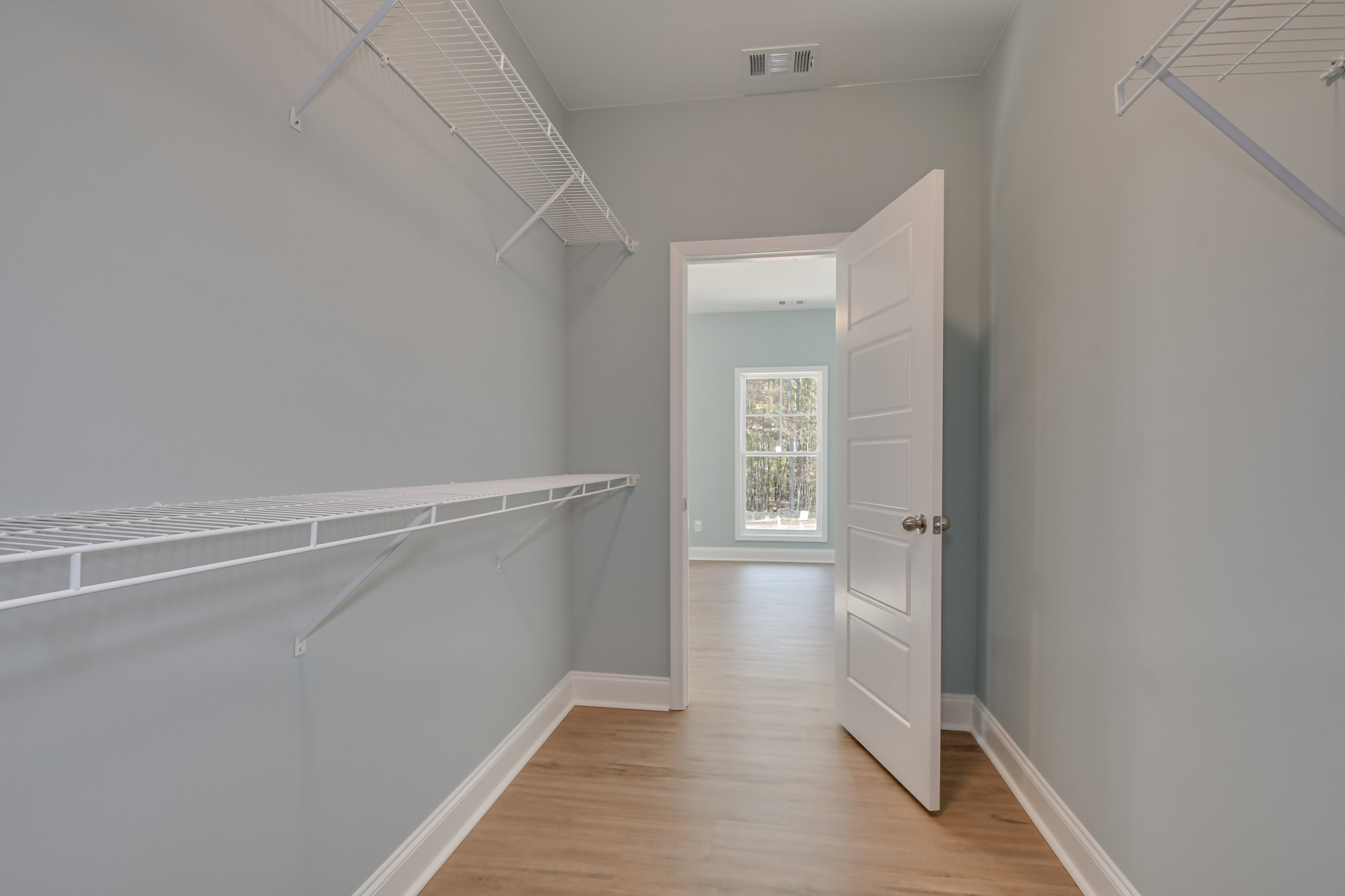Open white door with silver doorknob, wood floor with white trim, built-in white shelf on wall, window showing trees outside