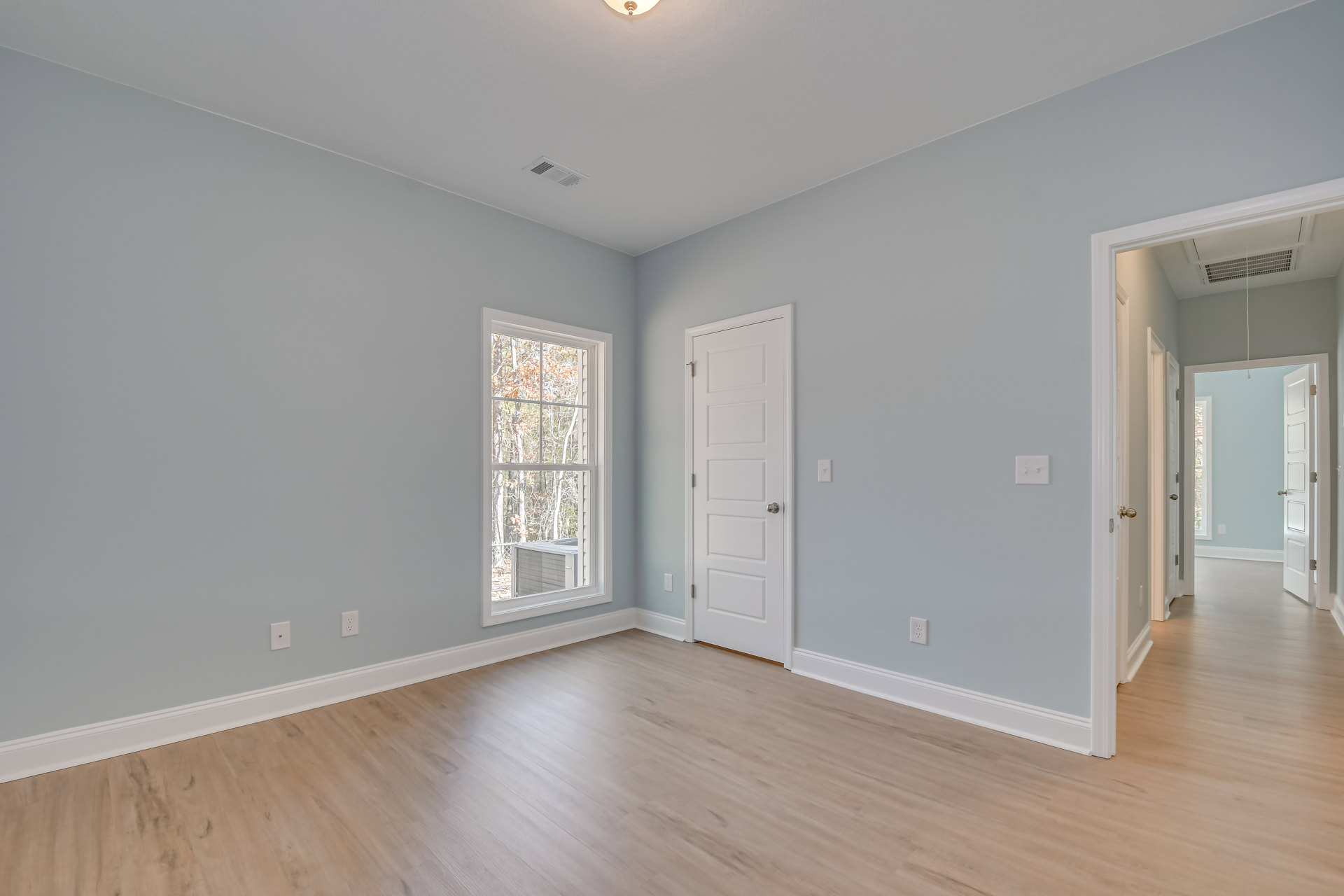 Room with light wood flooring, white walls, large window overlooking trees, white door with silver knob, ceiling light fixture, and decorative molding.