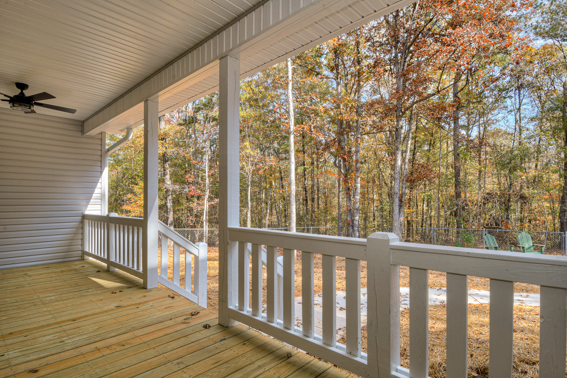Covered porch with white railing, green outdoor chair, ceiling fan with light, trees and autumn foliage in background, close-up of porch post, grey rectangular planter with straw.
