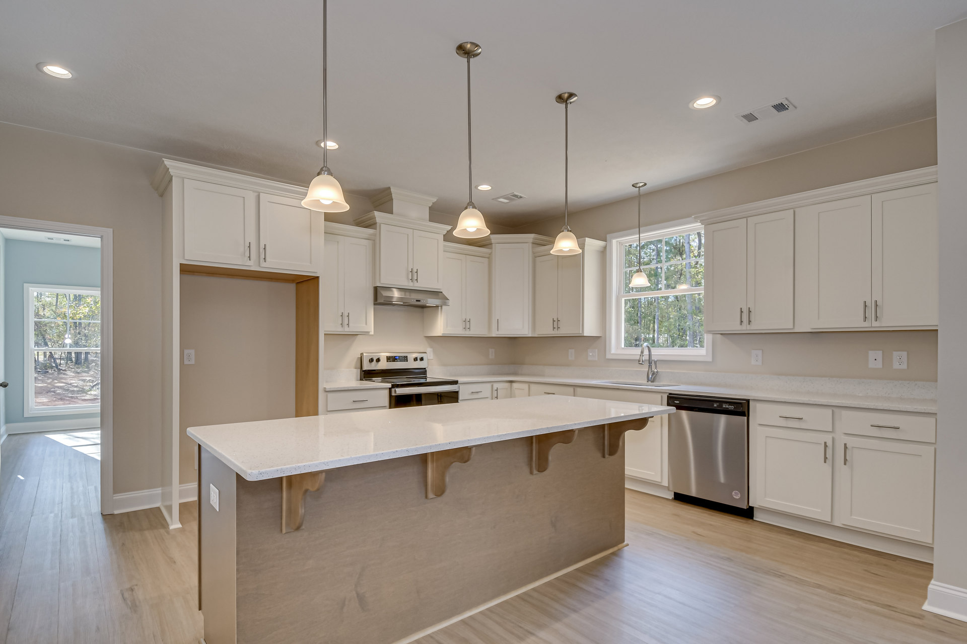 Kitchen featuring a spacious island with quartz countertop, stainless steel dishwasher, white cabinetry, undermount sink, pendant lighting, and large window overlooking trees.