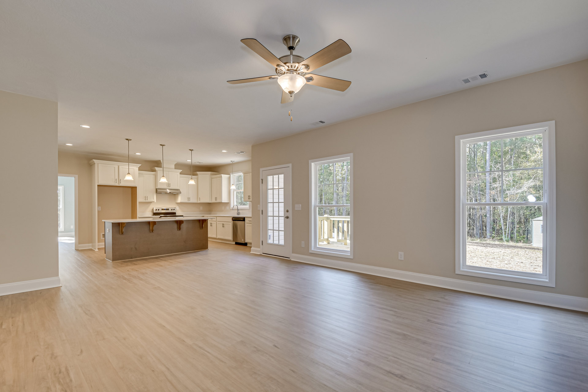 Open floor plan featuring wood flooring, kitchen with white cabinetry, dining area, ceiling fan with light fixture, large windows overlooking trees, and a white glass-paneled door.