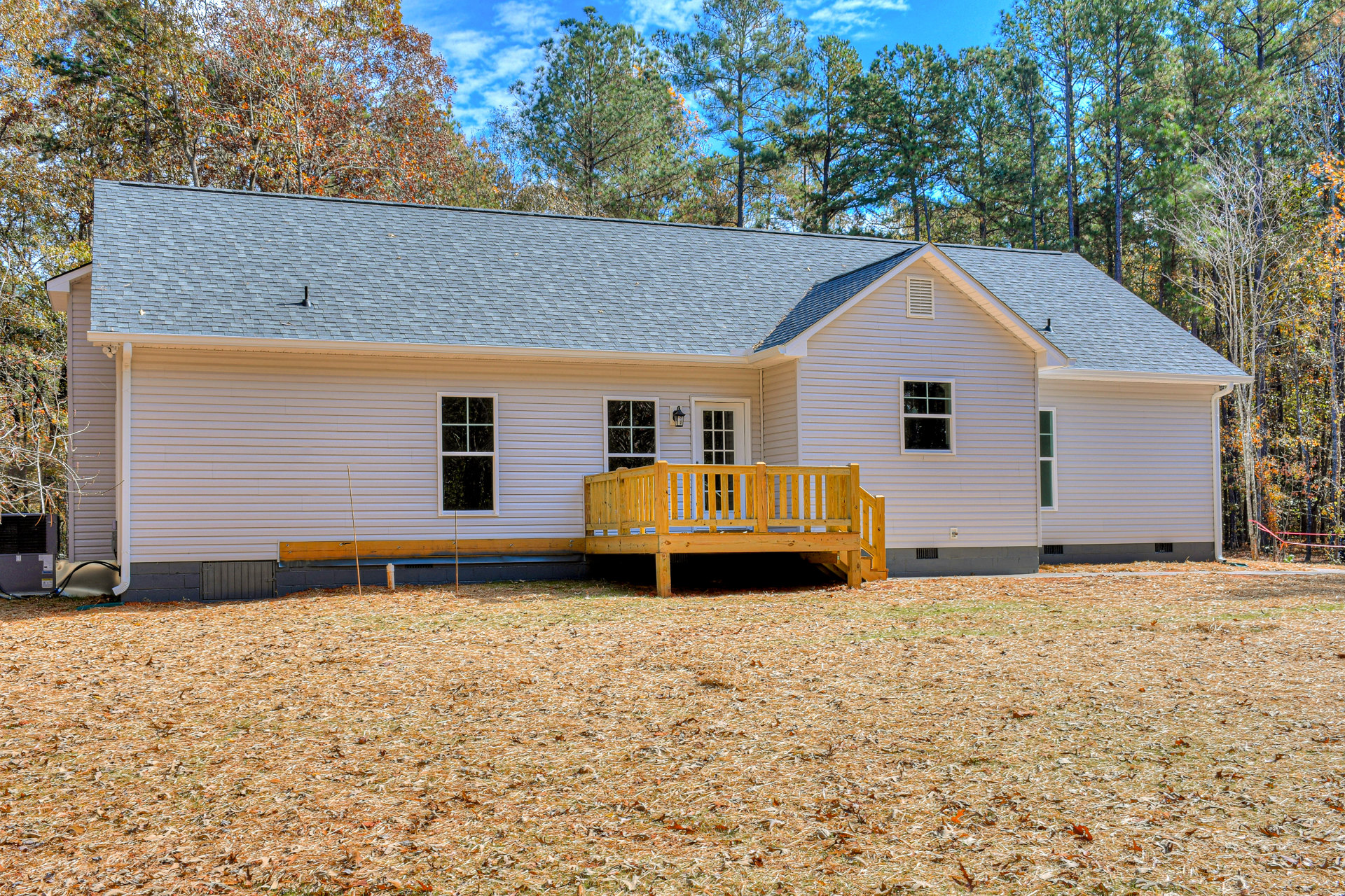 Wooden deck with white railing overlooking a yard covered in brown leaves, large window with white frame, gray siding exterior, mature trees in background