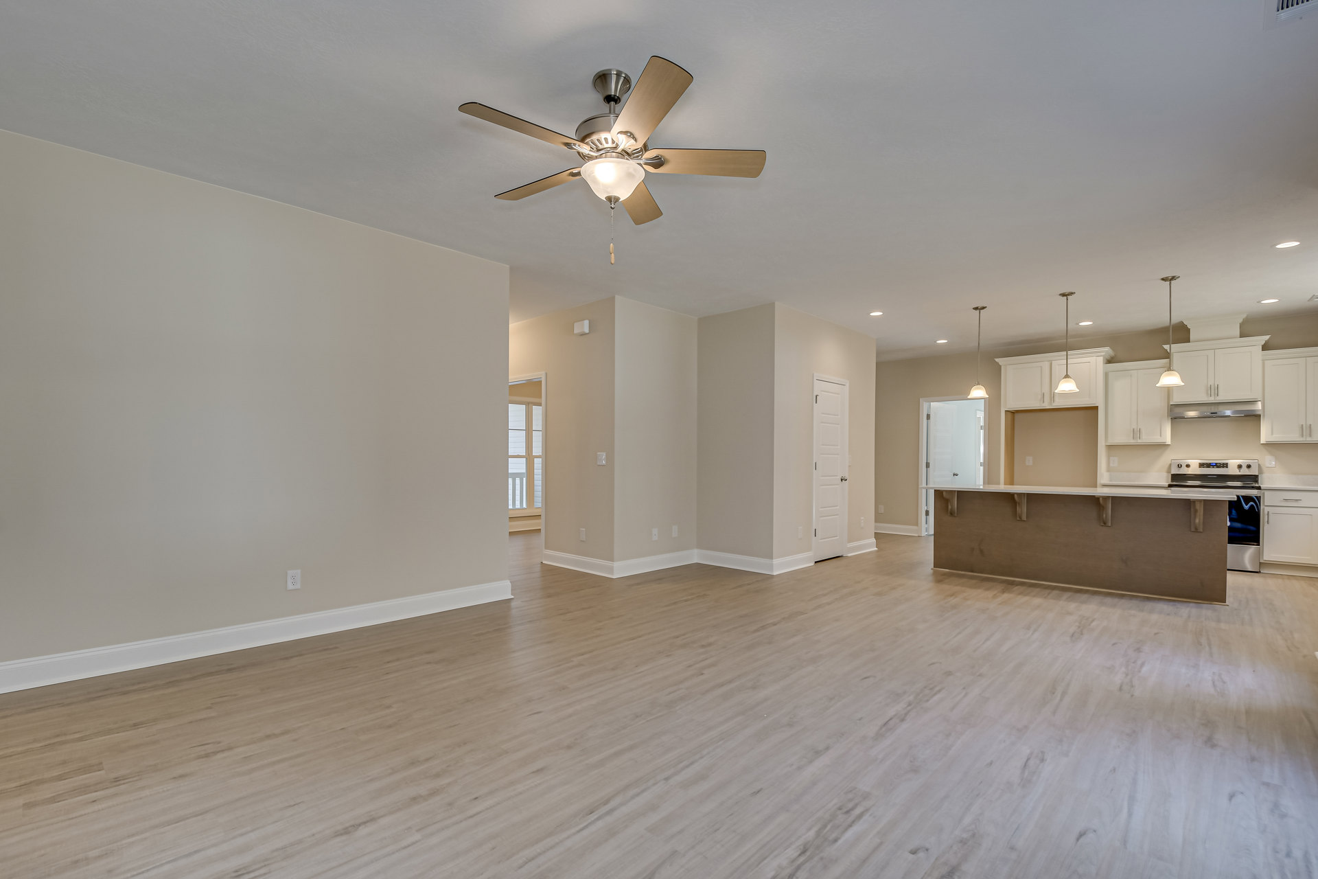 Living room with hardwood floors, white walls, ceiling fan with light fixture, open view into kitchen area, white door with silver handle