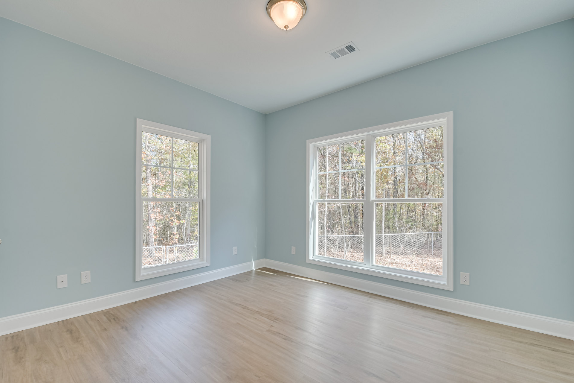 Light blue painted wall, two windows with tree views, wood laminate flooring, white ceiling with vent and recessed light, simple molding trim
