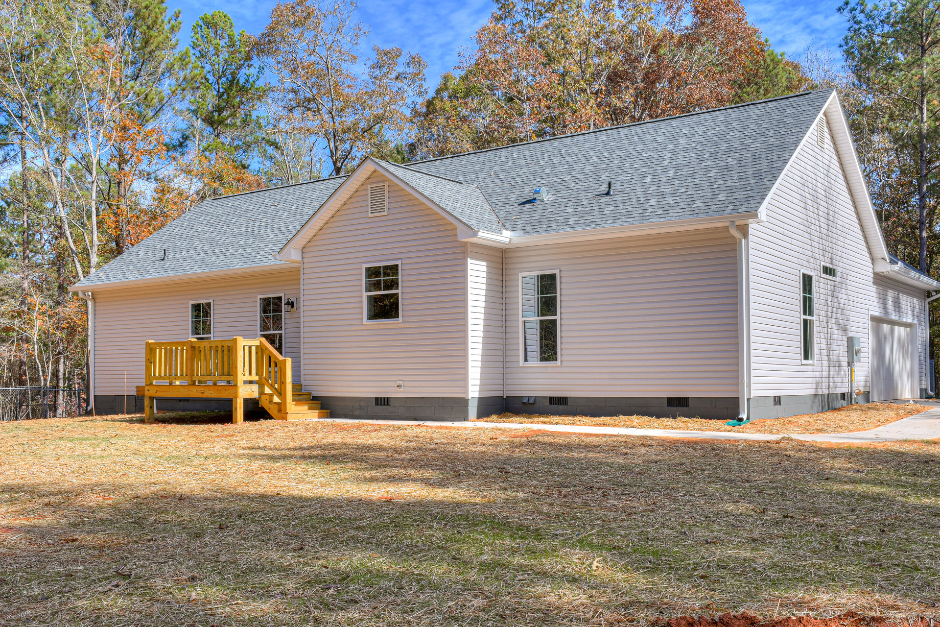 Two-story house with gray siding, white-framed windows, wooden deck with railing, grassy yard, and mature trees in the background