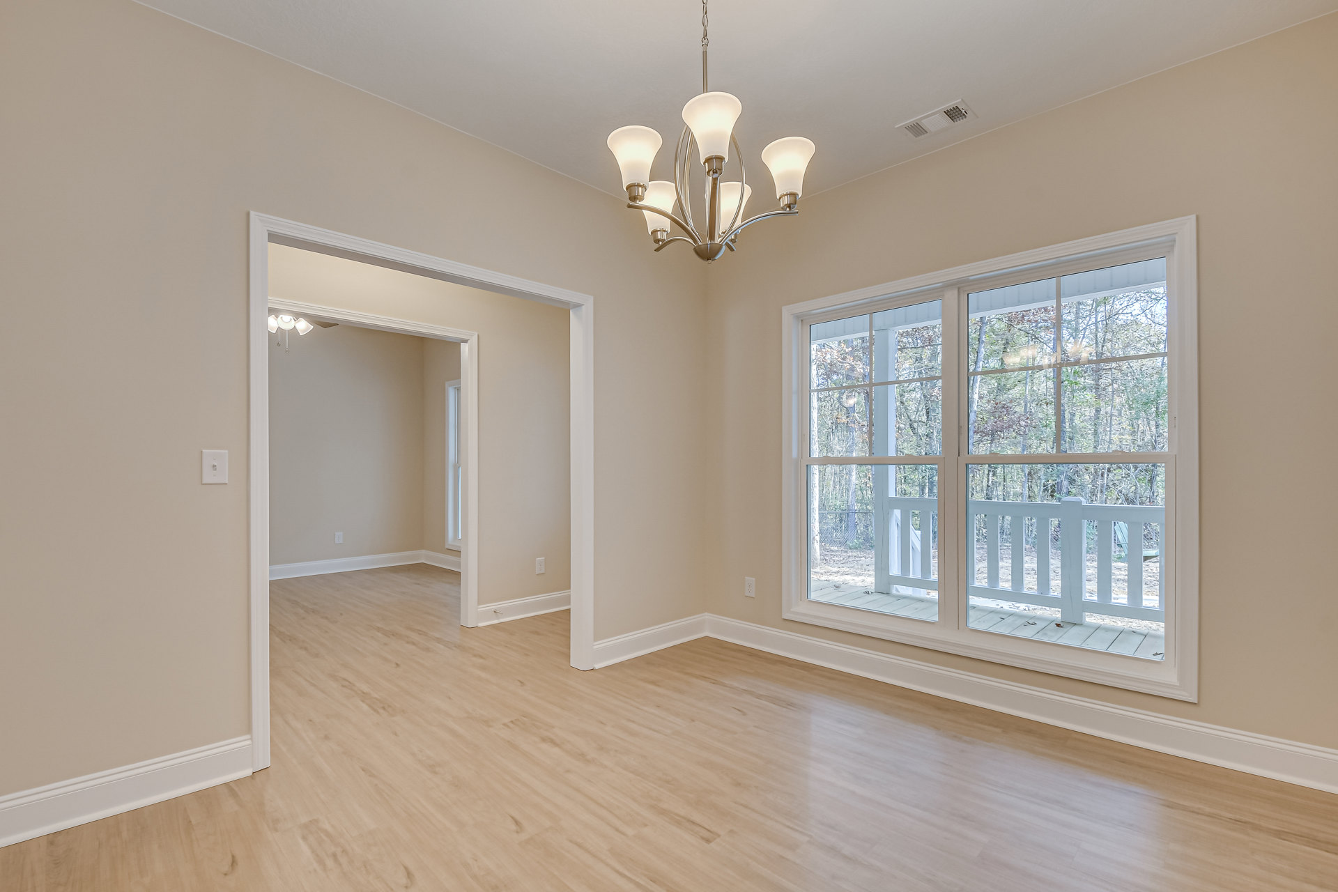 Open living space with wide plank wood flooring, ornate chandelier, large windows overlooking deck and trees, white walls with crown molding