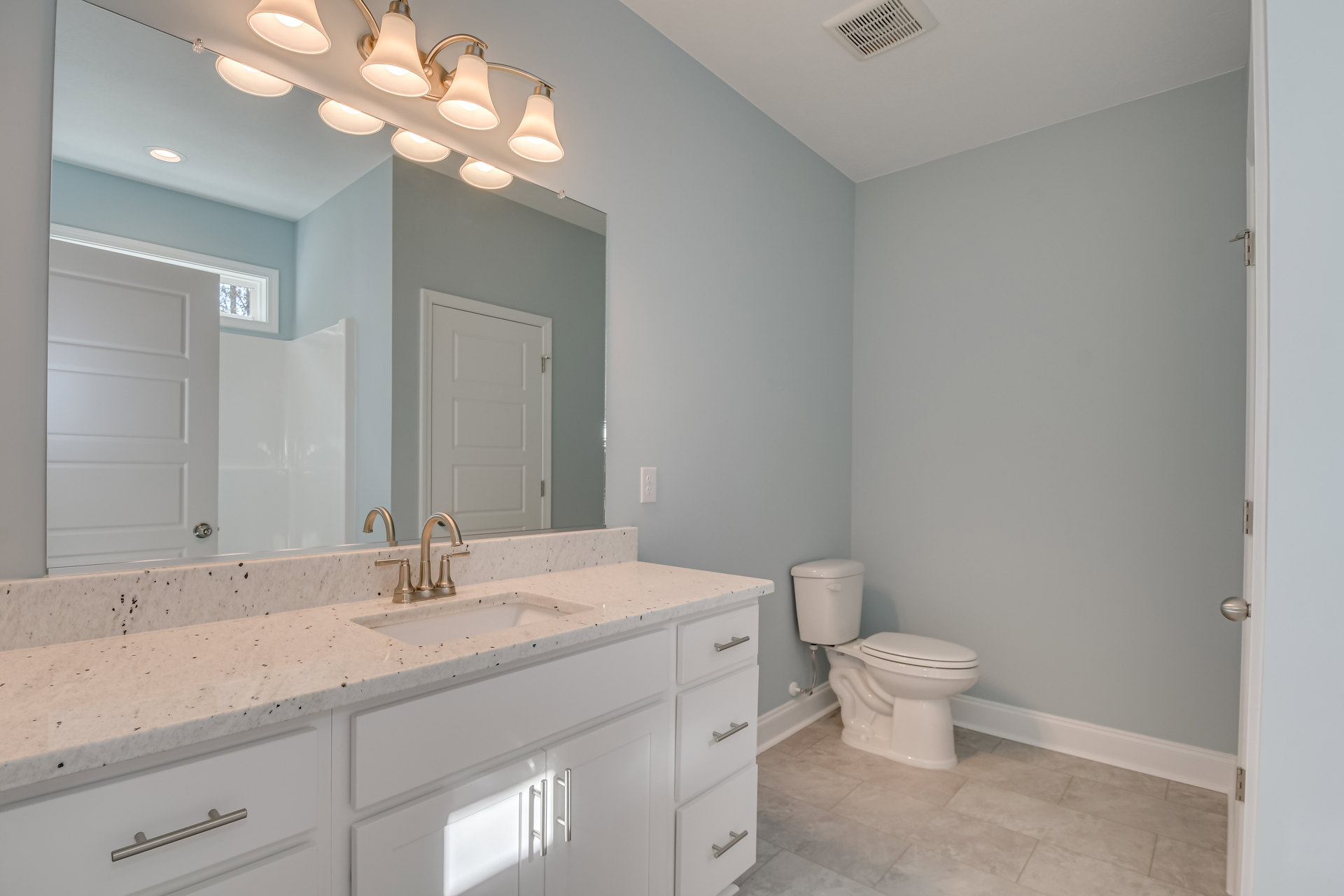 Marble countertop vanity with undermount sink, white toilet, light gray tile floor, white cabinetry, silver faucet, multi-bulb wall light fixture, white door with silver handle