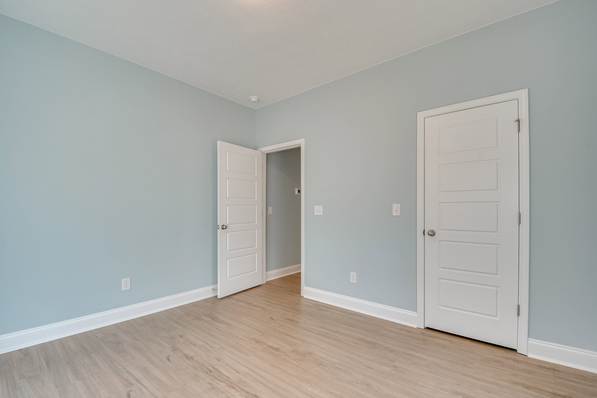 Room with two white doors, silver door knobs, wood laminate flooring, white baseboard trim, white cabinet, and smoke detector on ceiling