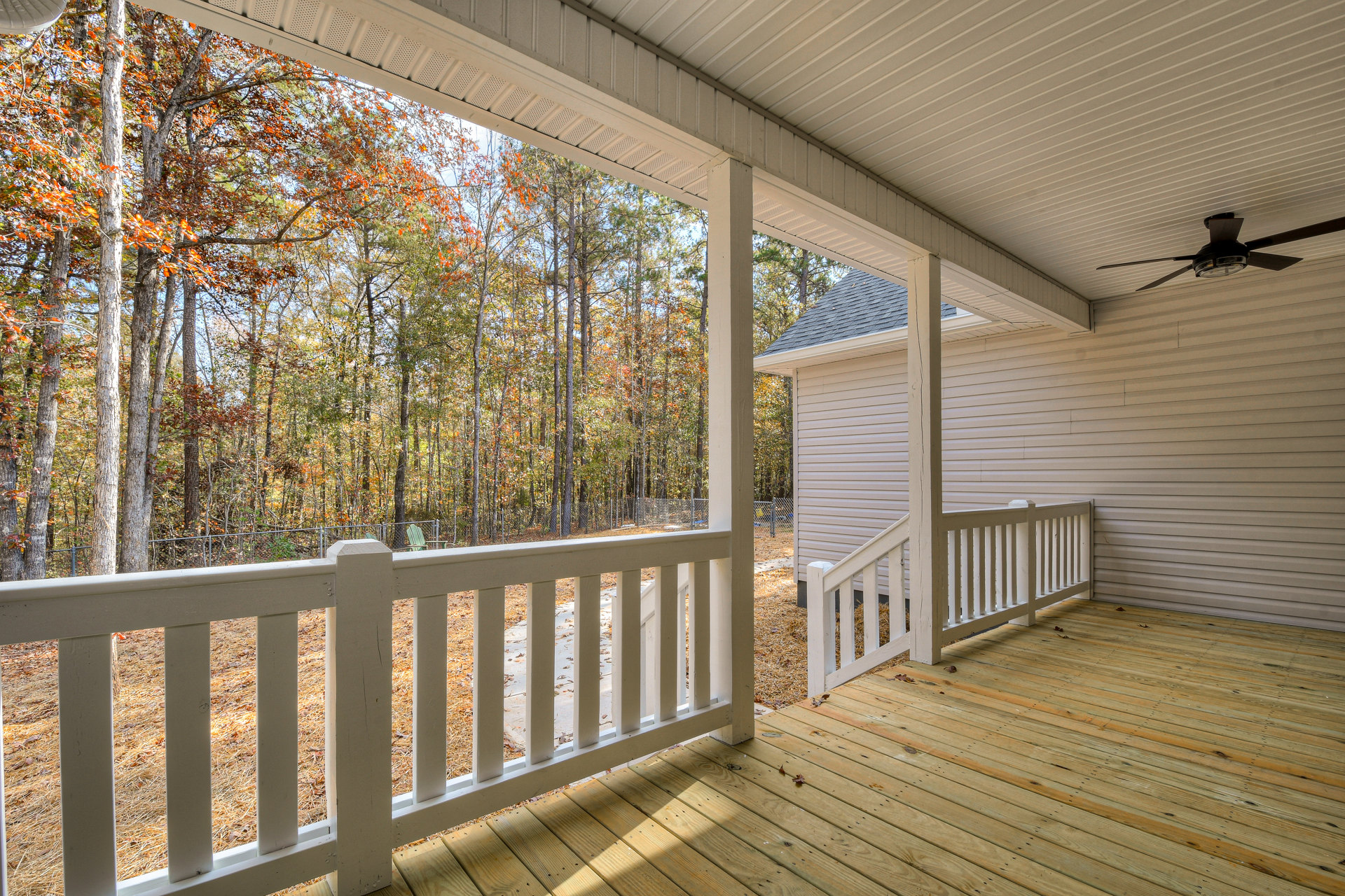Deck with white railing and balusters, covered porch area, autumn trees in background, white exterior siding, large windows, outdoor ceiling.