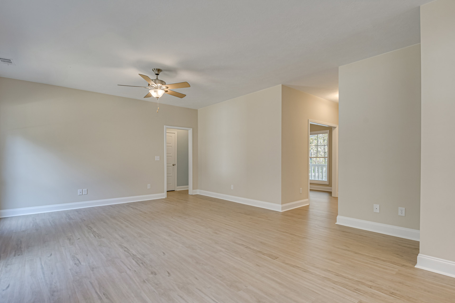 Ceiling fan with light fixture above hardwood floor, white baseboard trim, window showing trees outside, white door with silver knob