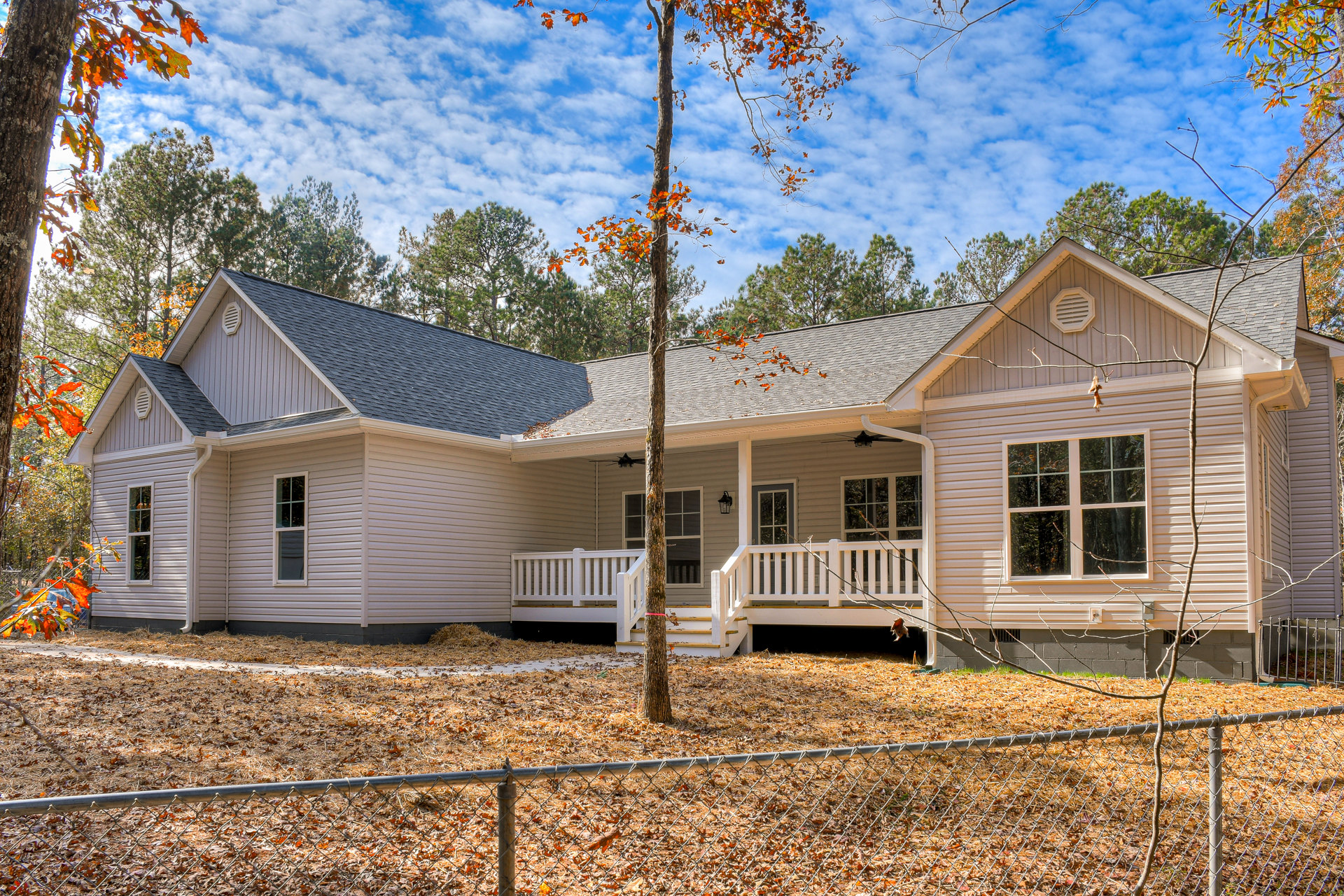 Two-story home with light siding, white porch railing, and large windows reflecting trees; wooden fence and mature trees with orange leaves in the landscaped yard.