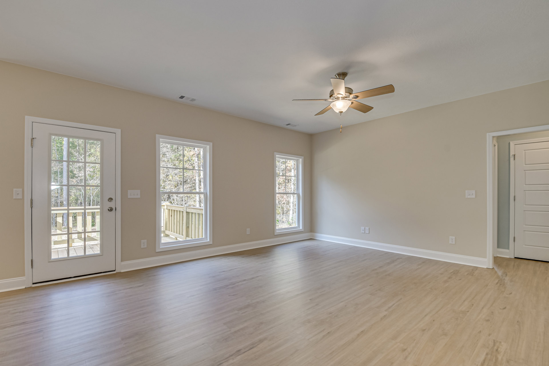 Ceiling fan with light fixture above wood flooring, glass-paneled door, and windows overlooking trees, wood railing visible outside