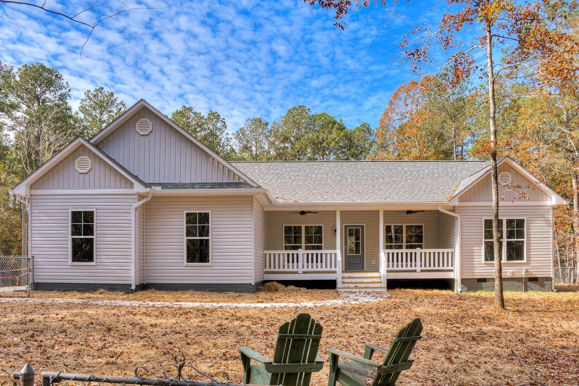 Two-story house with white siding, large front window, and glass-paneled door; green wooden lawn chair sits on grassy lawn bordered by leafy trees under partly cloudy sky.