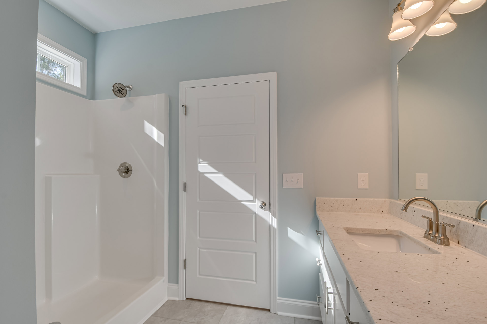 Bathroom with white paneled door and silver knob, rectangular sink with chrome faucet, tiled walls, ceiling-mounted light fixtures, and modern showerhead.