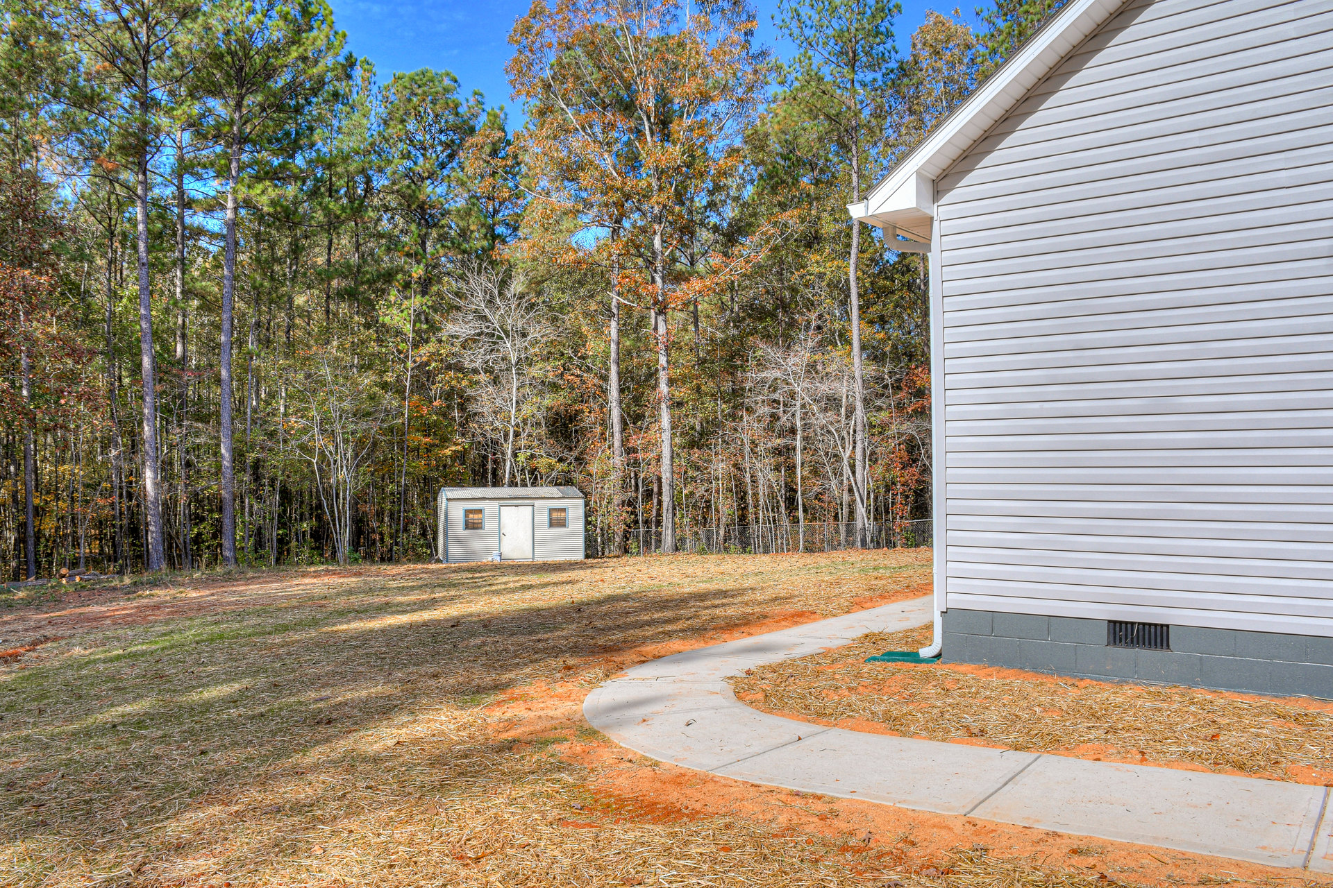 White house with a concrete path bordered by grass and dirt, orange-leaved deciduous tree, white shed with door, trees in the background, autumn sky