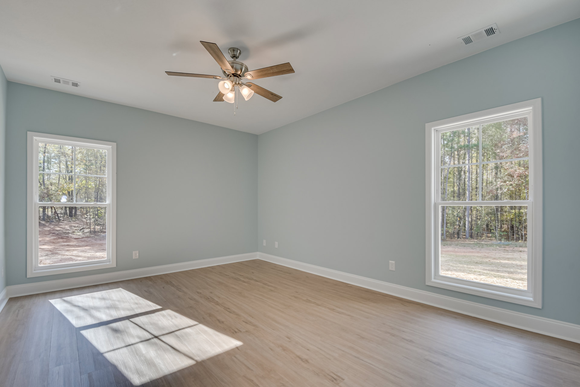 Ceiling fan with light fixture, wood flooring with white trim, large windows showing trees outside, smooth white walls and ceiling