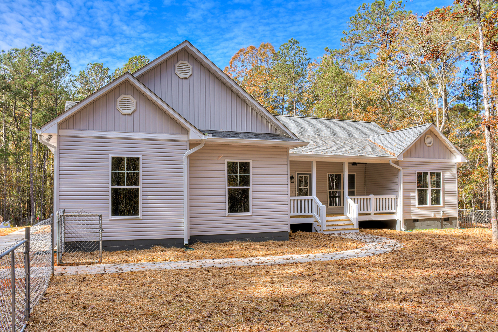 Two-story home with light siding, white-framed windows reflecting trees, paved driveway, chain link fence with metal gate, mature trees in the background, white vent on exterior