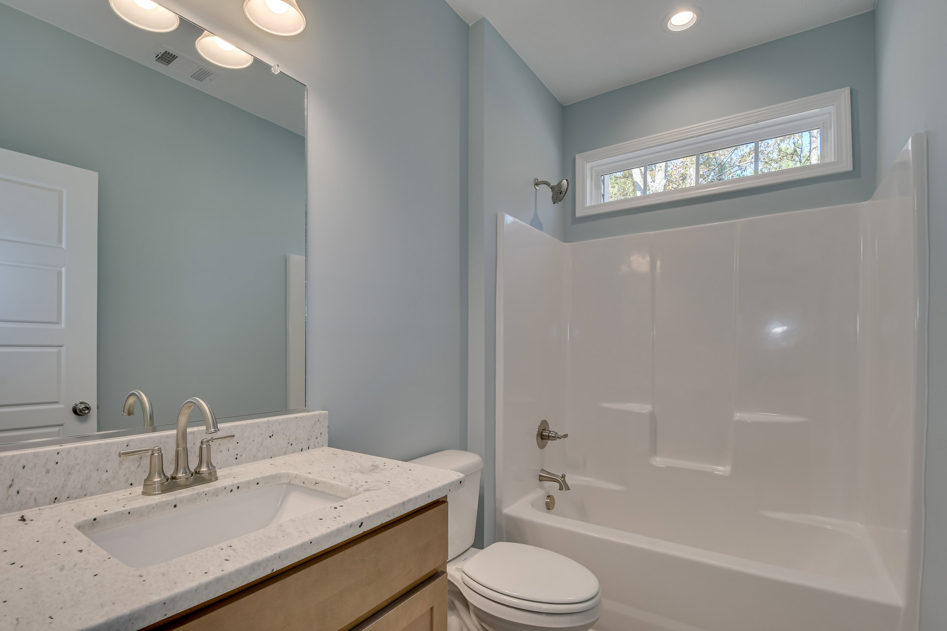 Bathroom with white porcelain sink and chrome faucet, white toilet, rectangular bathtub, light-colored tile walls, large window showing green trees outside