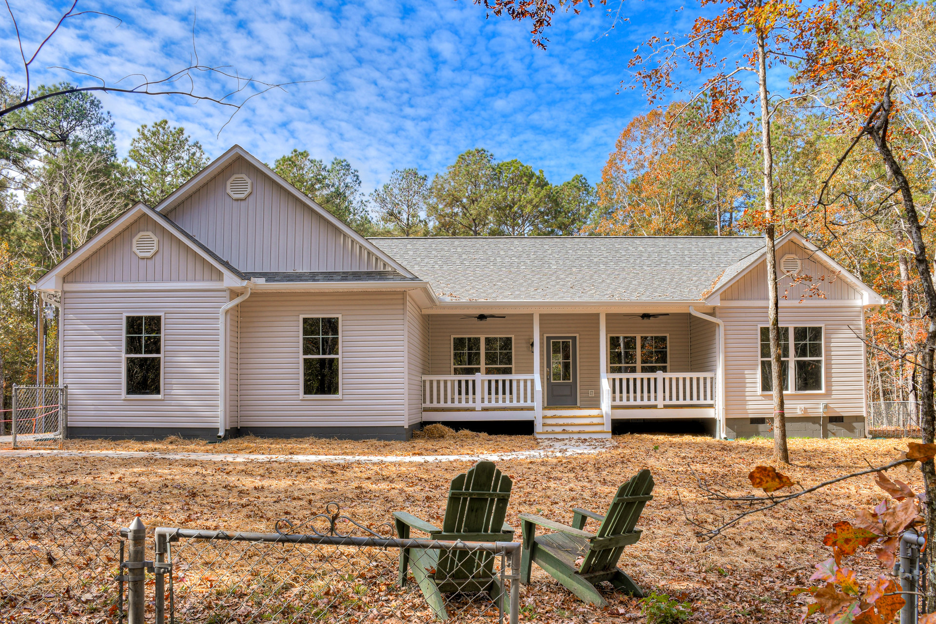 Green wooden lawn chair on fallen leaves beside white house with window, vent, and door with glass panel; white-framed window and partial fence visible; autumn trees in background.