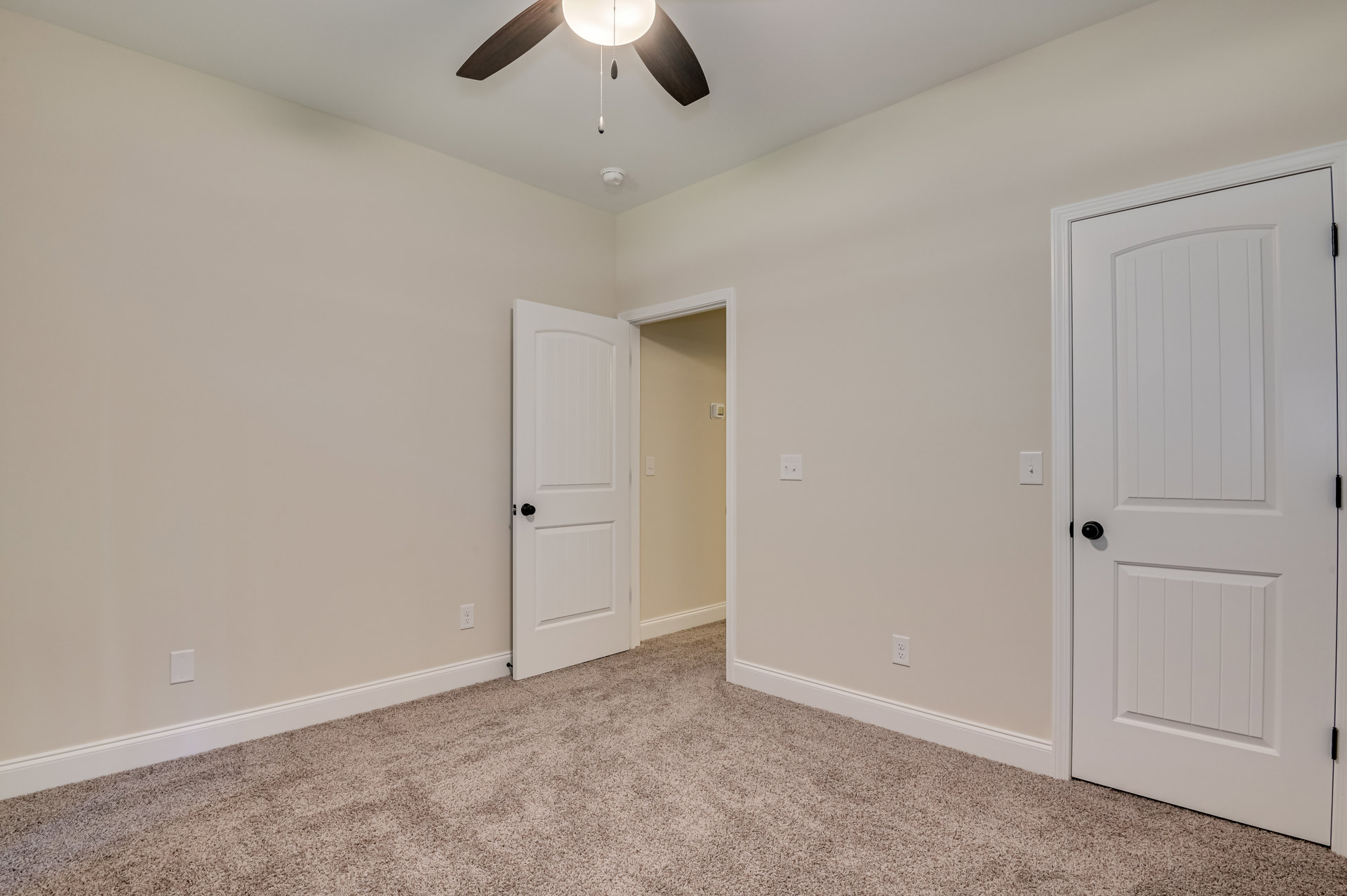 Carpeted room with white trim, ceiling fan with light fixture, two white doors featuring black knobs, neutral walls