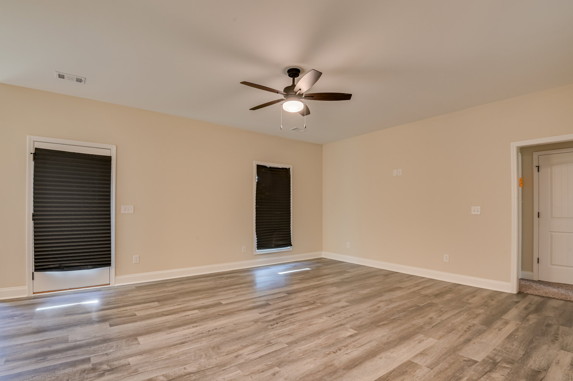 Ceiling fan with light fixture, hardwood flooring, white walls, window with black shade, white-framed door with illuminated light switch