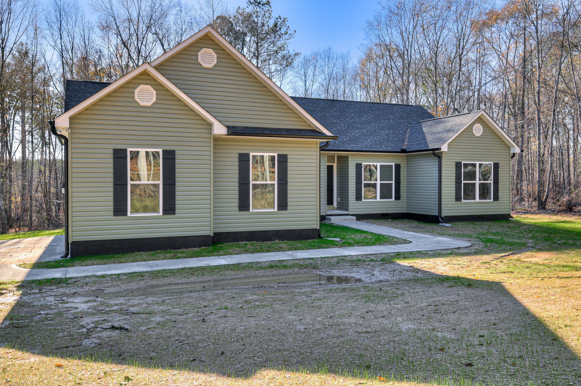 Modern home with light siding, large windows reflecting surrounding trees, white vent detail, attached garage with white door, concrete driveway bordered by grass, puddle near