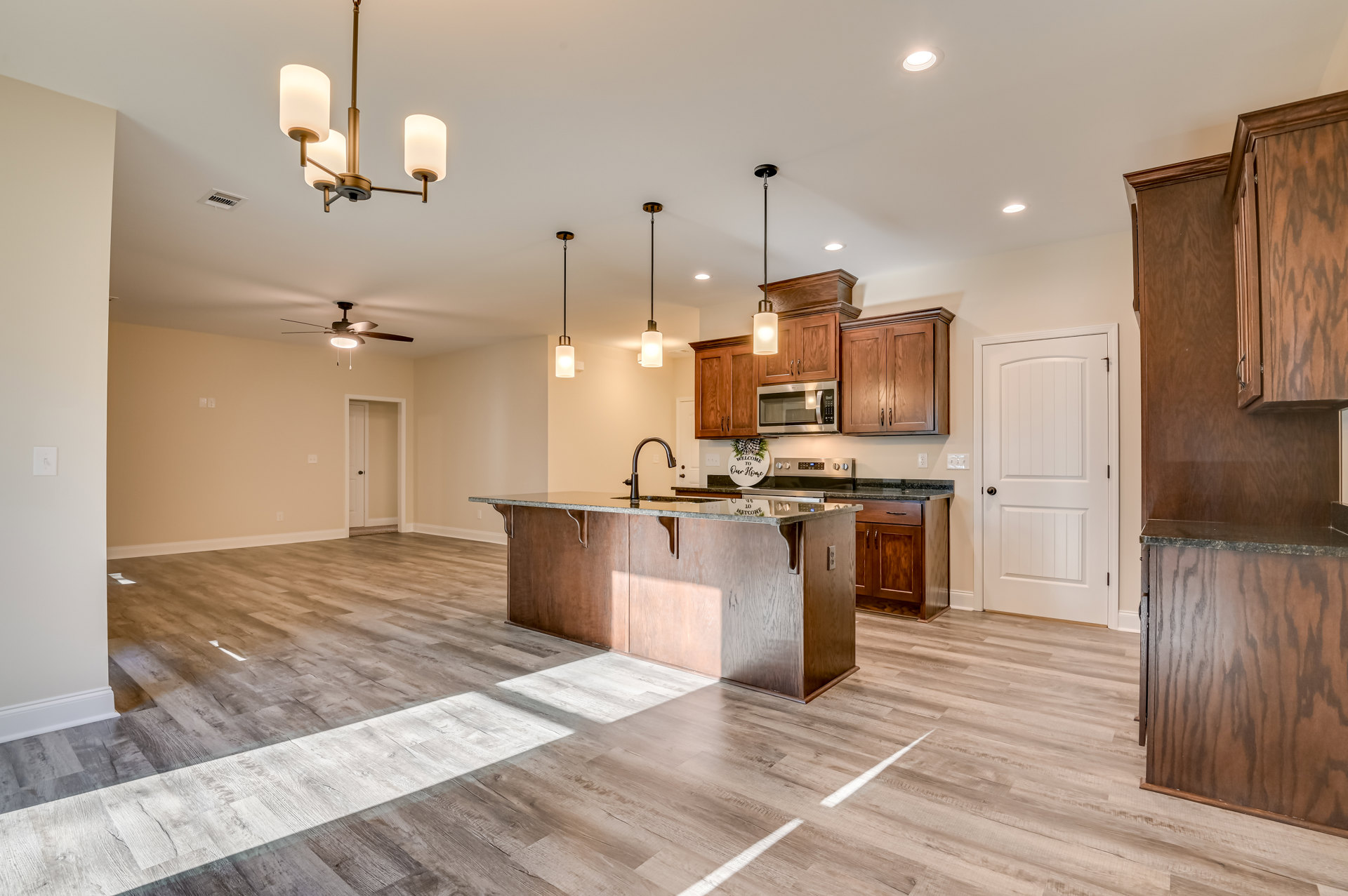 Open-concept kitchen and dining area with wood flooring, wooden cabinets topped with black countertops, open microwave door, white door with black knob, decorative sign with bow
