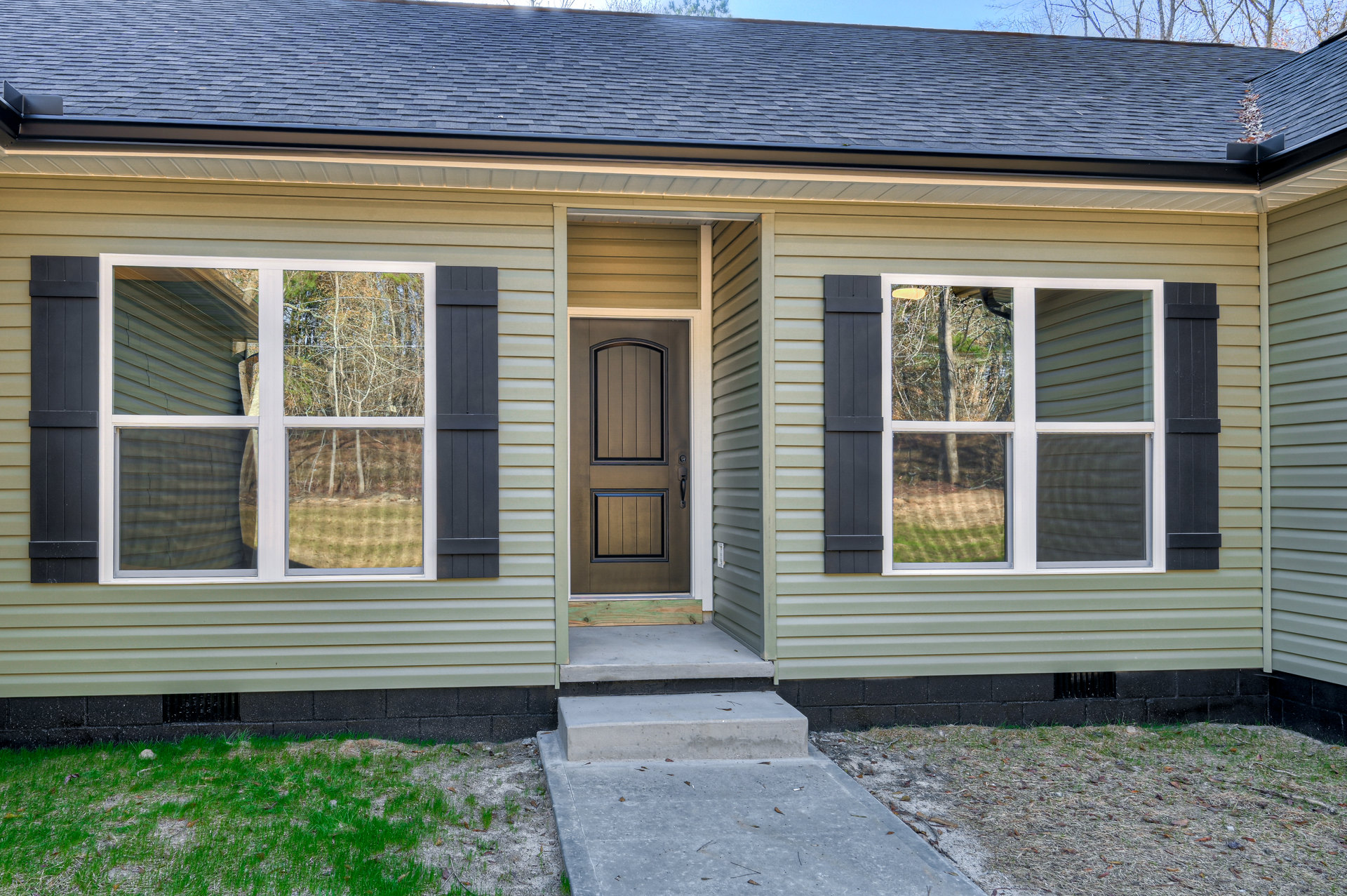 Modern home exterior with light-colored siding, large windows reflecting trees and grass, wooden front door, concrete porch, and landscaped greenery