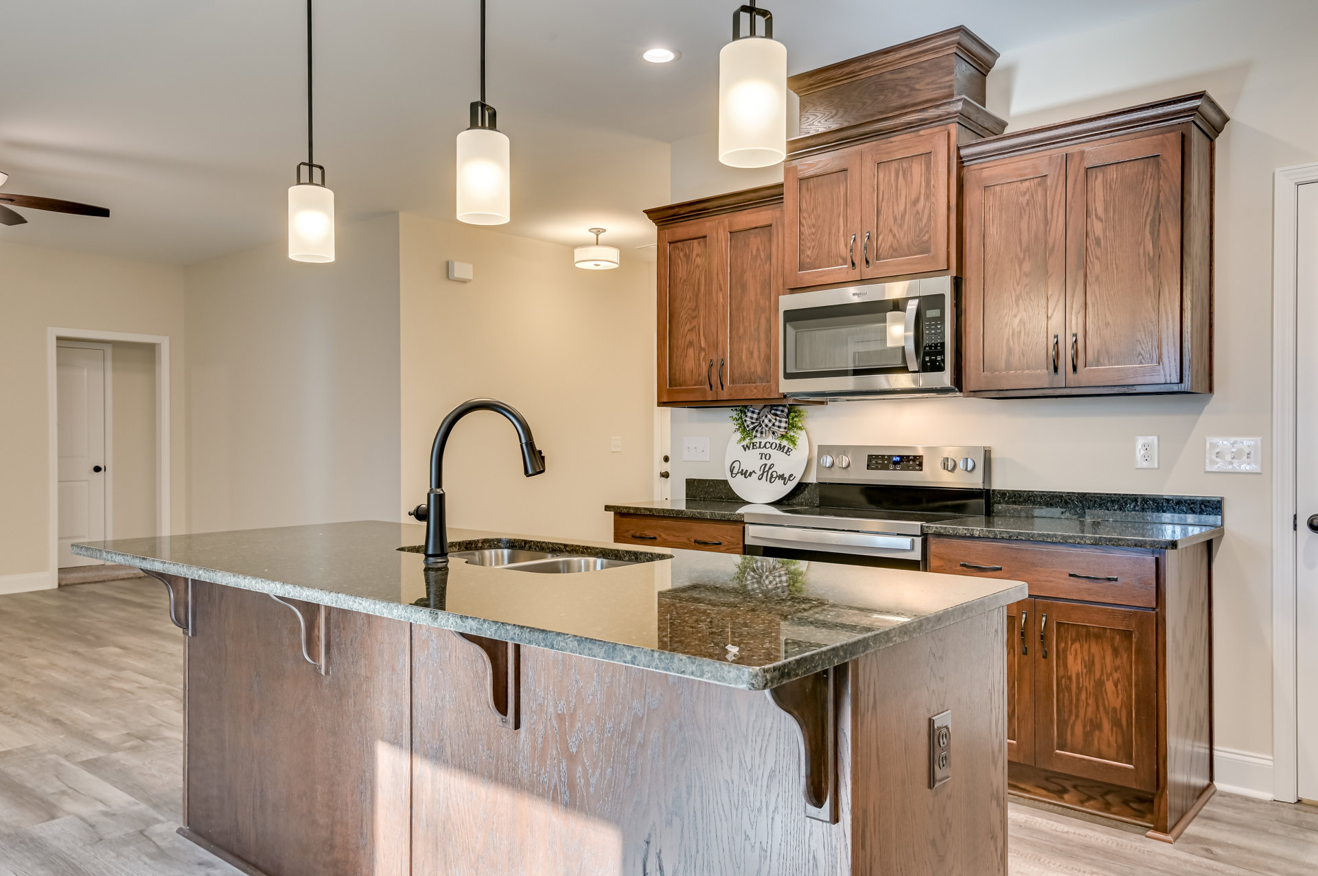 Granite countertop kitchen with stainless steel stove, built-in microwave, undermount sink, white cabinetry, black pendant lamp, and decorative sign with bow.