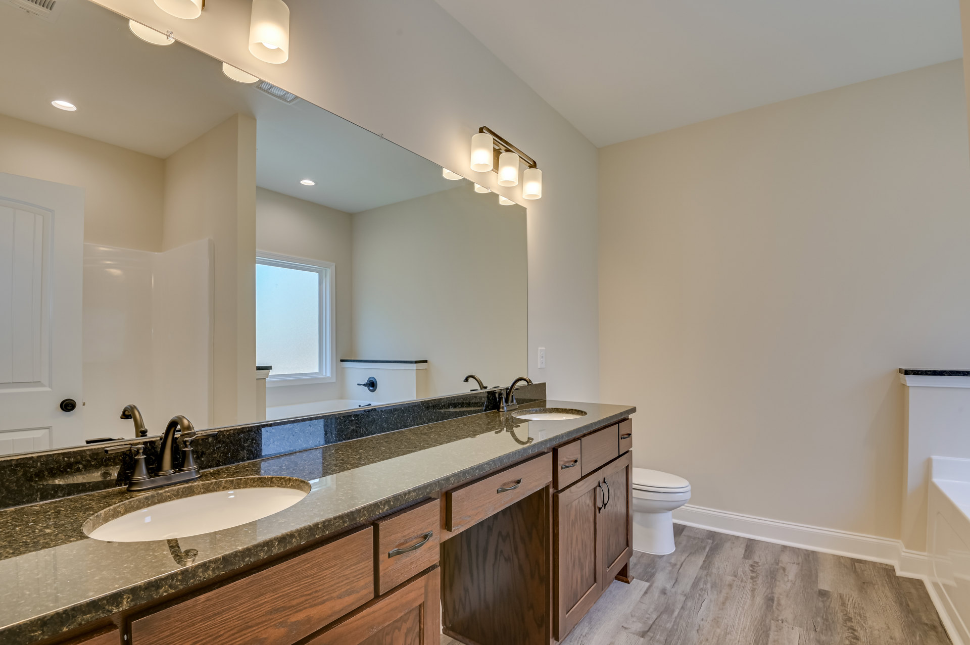 Bathroom with marble countertop double sinks, wide framed mirror, white cabinetry, chrome faucets, tiled floor, window showing blue sky, closed white toilet.