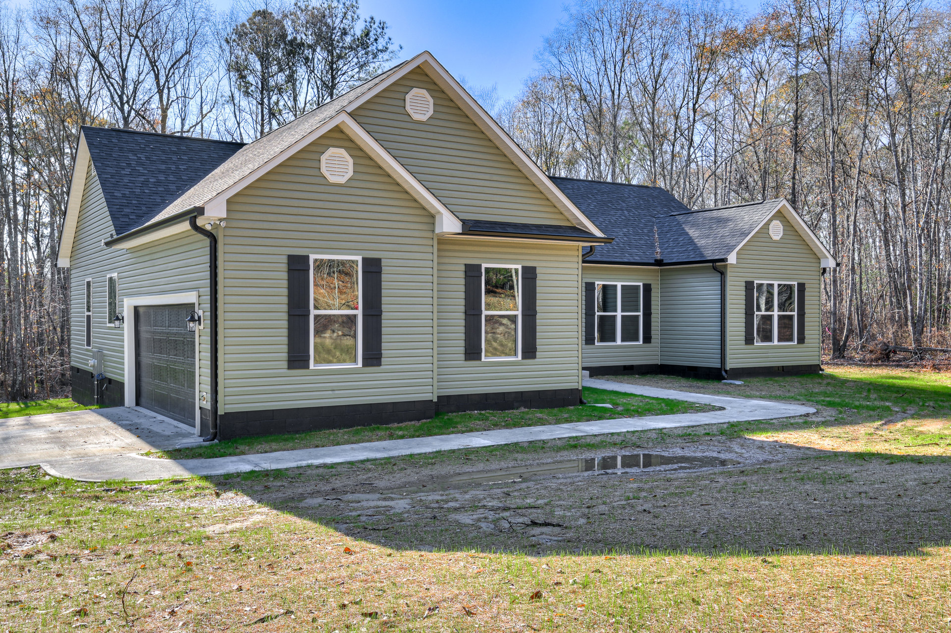 Two-story house with light-colored siding, white-framed windows, and a paved driveway; mature trees and blue sky in the background; one window shows broken glass and tree