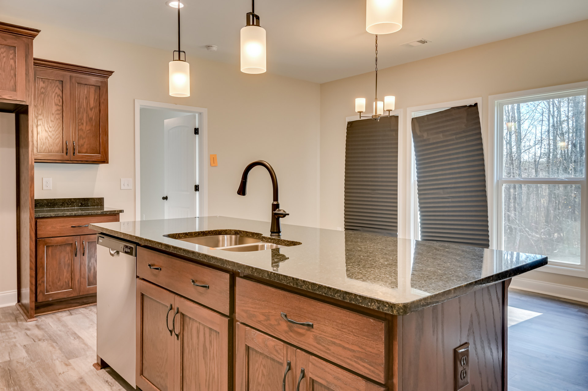 Modern kitchen with two stainless steel sinks set in a light stone countertop, white cabinetry, grey and black window blinds, large window overlooking trees, rectangular ceiling